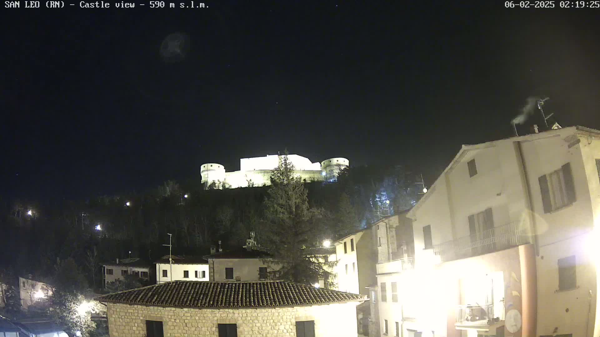 A nighttime view of a hilltop castle illuminated against a dark, clear sky, overlooking a village with buildings lit up.