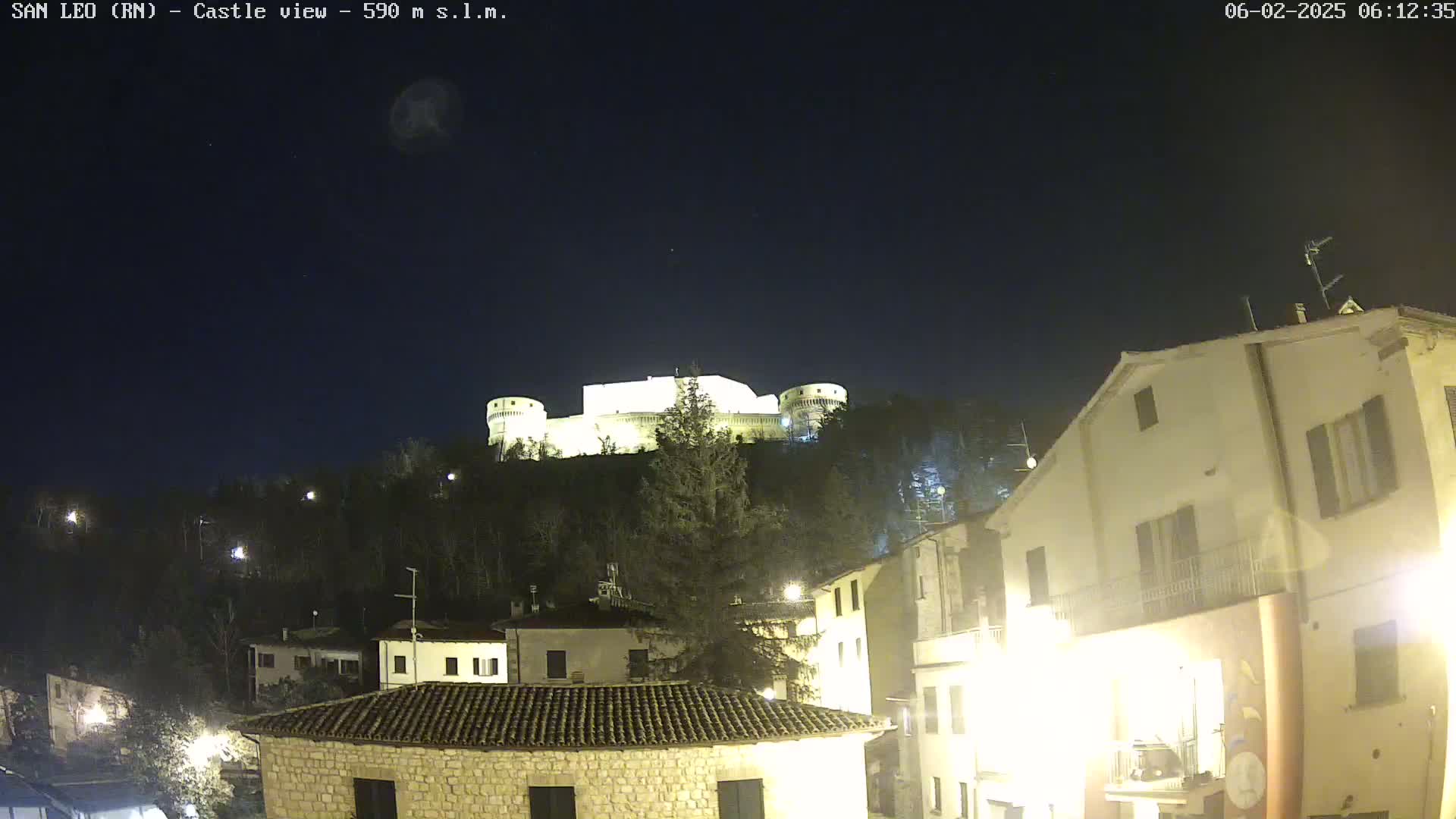 A nighttime view of a hilltop castle illuminated against a dark, clear sky, overlooking a town with buildings lit by streetlights.