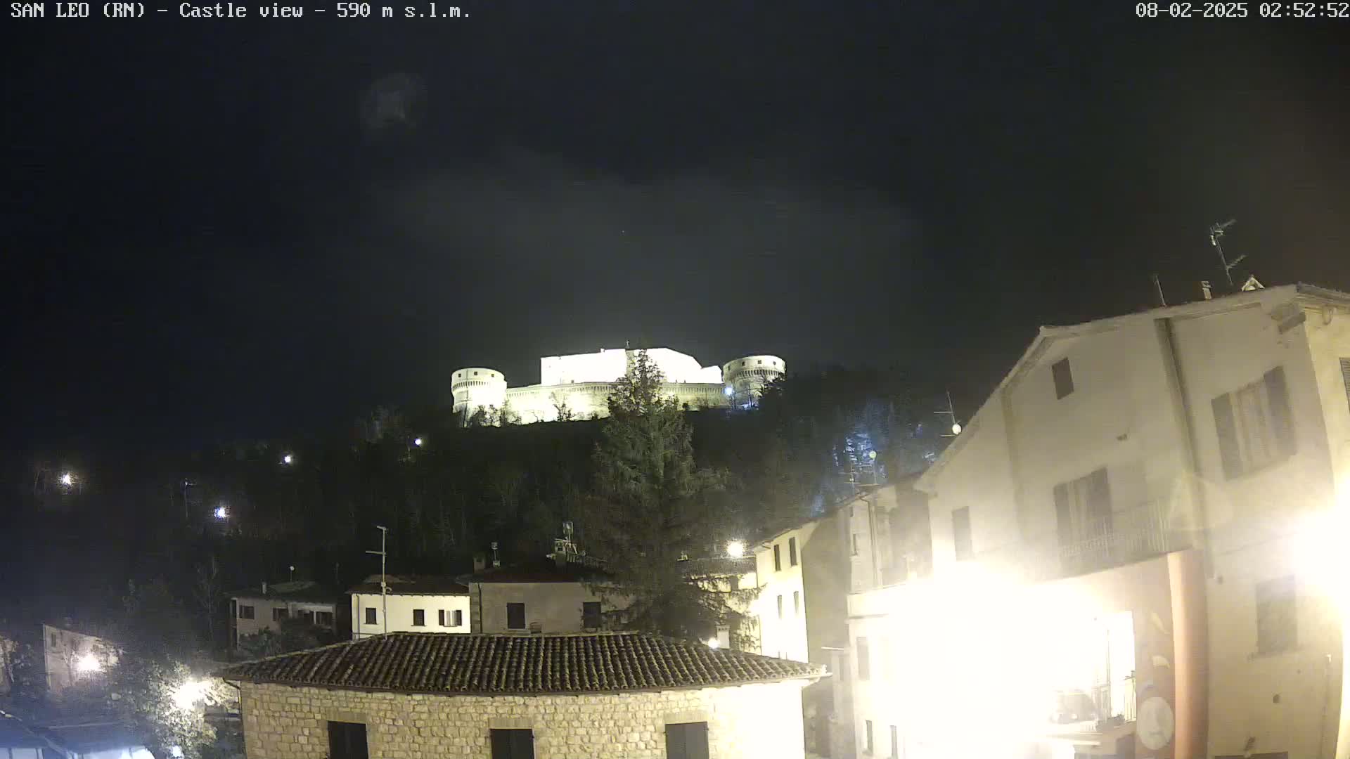 A nighttime view of a hilltop castle illuminated against a dark, clear sky, overlooking a village with buildings lit by streetlights.