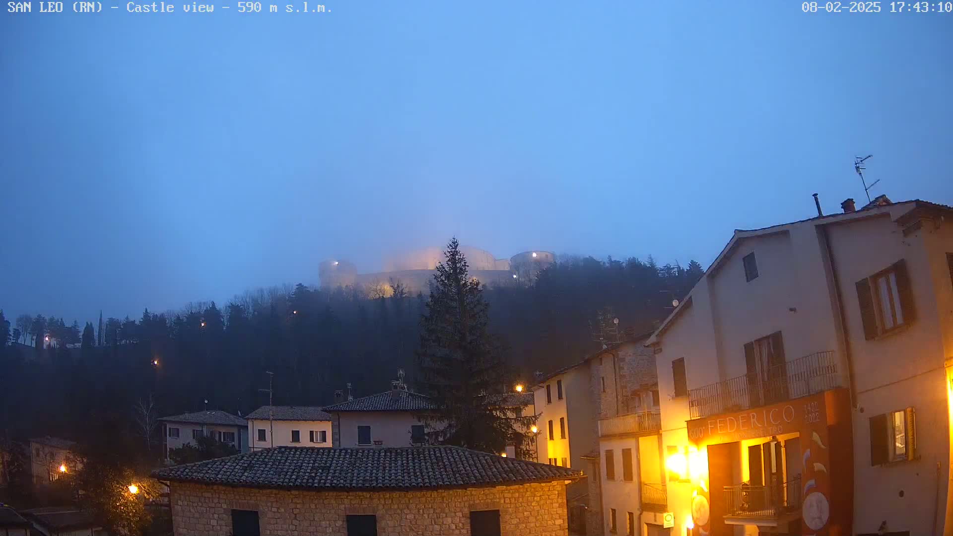 A hilltop castle is partially obscured by fog at dusk, overlooking a town with buildings lit by streetlights.