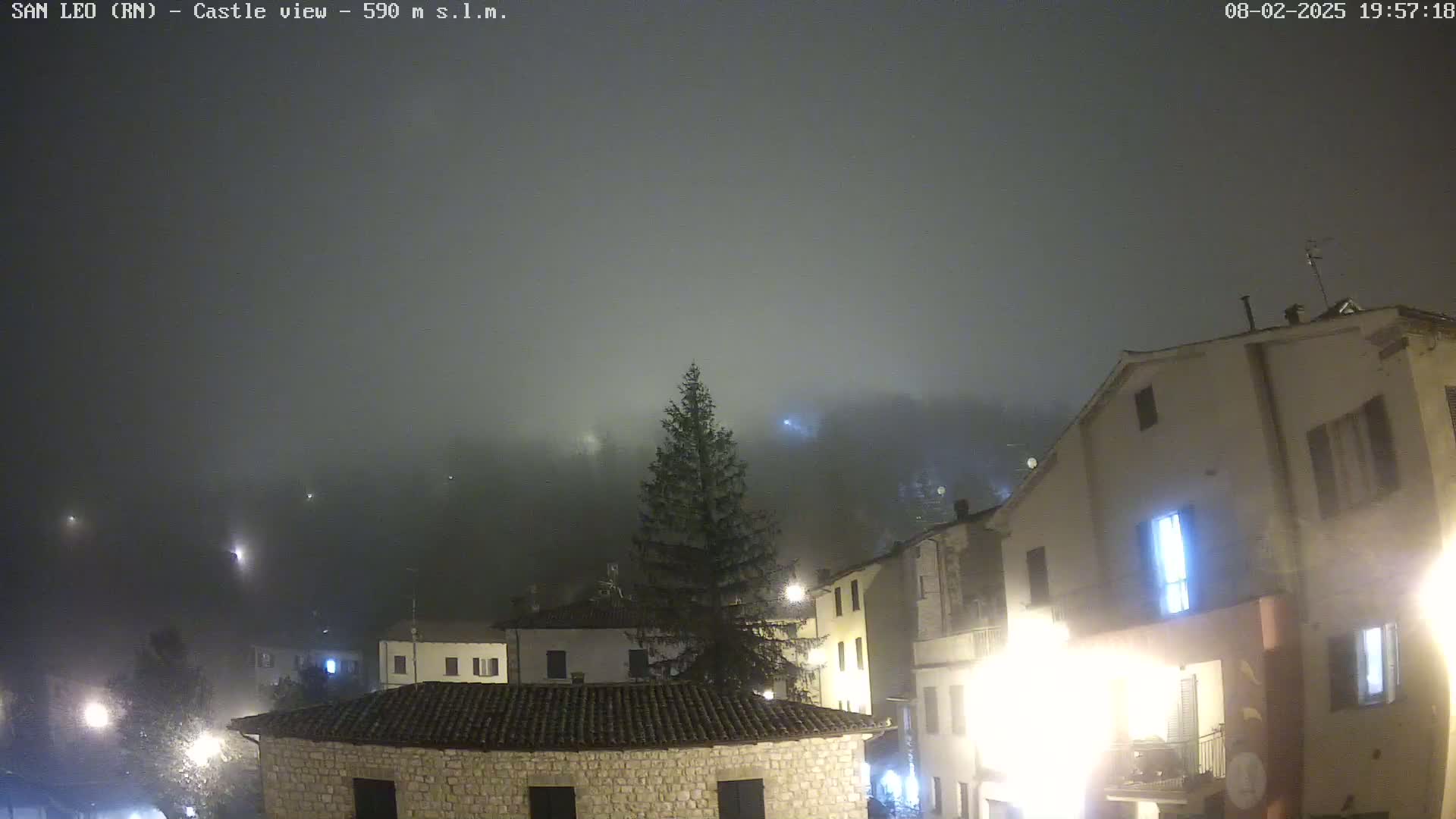 A foggy night scene shows a village of stone buildings lit by streetlights, with a tall evergreen tree in the foreground.