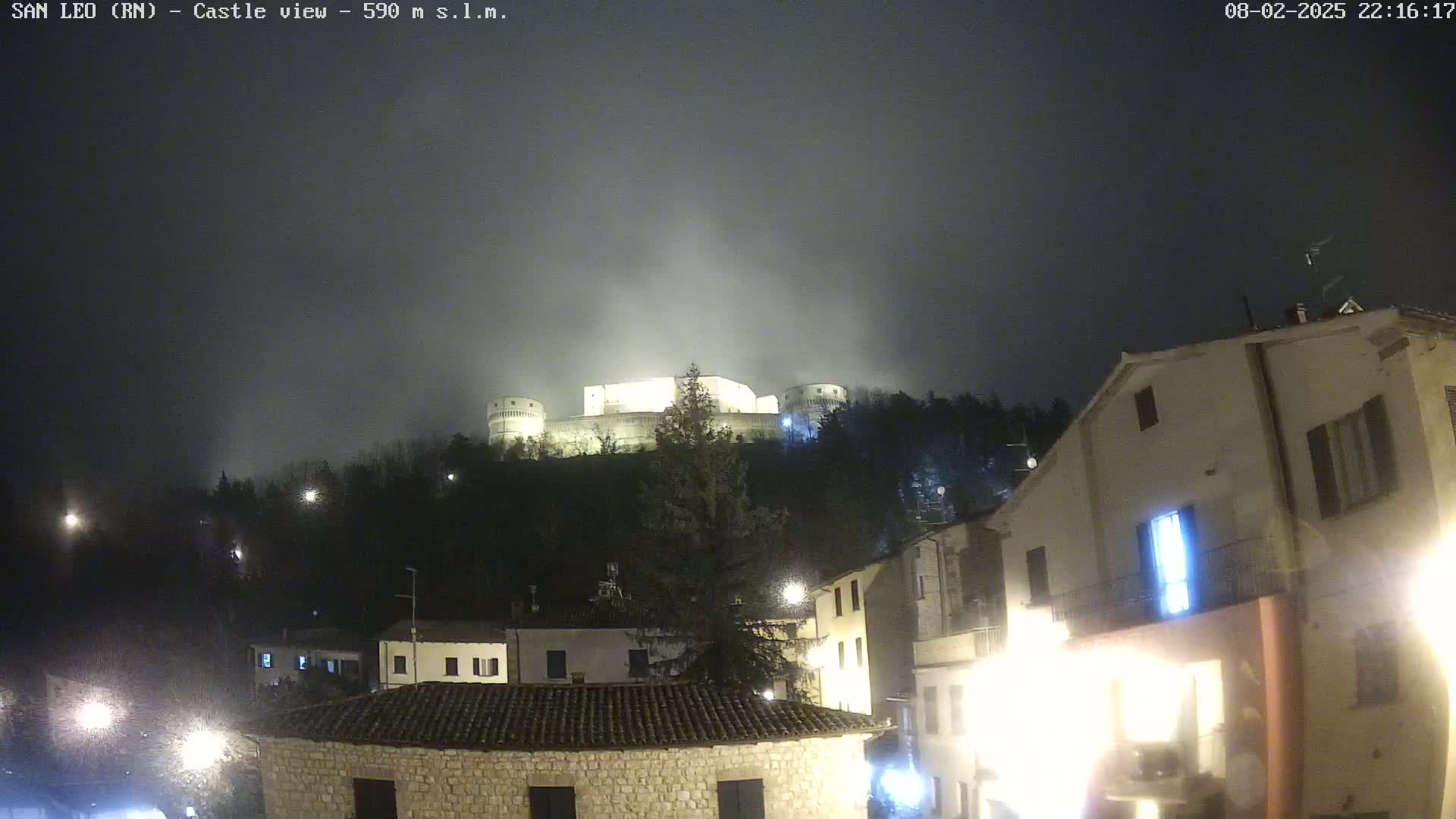 A nighttime, foggy view of a hilltop castle illuminated, overlooking a village with buildings and streetlights.