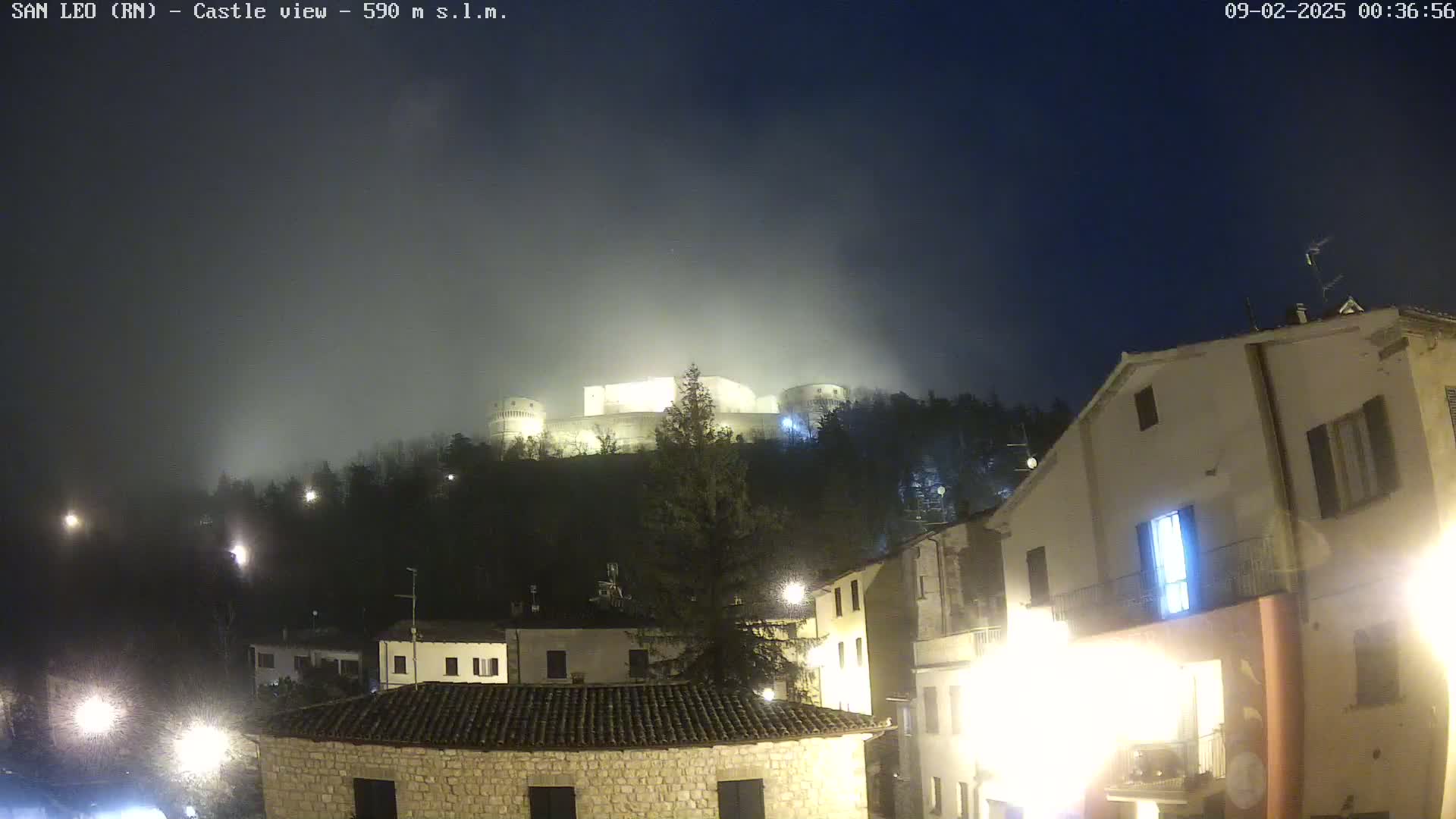 A fog-shrouded castle sits atop a hill overlooking a town at night, illuminated by streetlights.