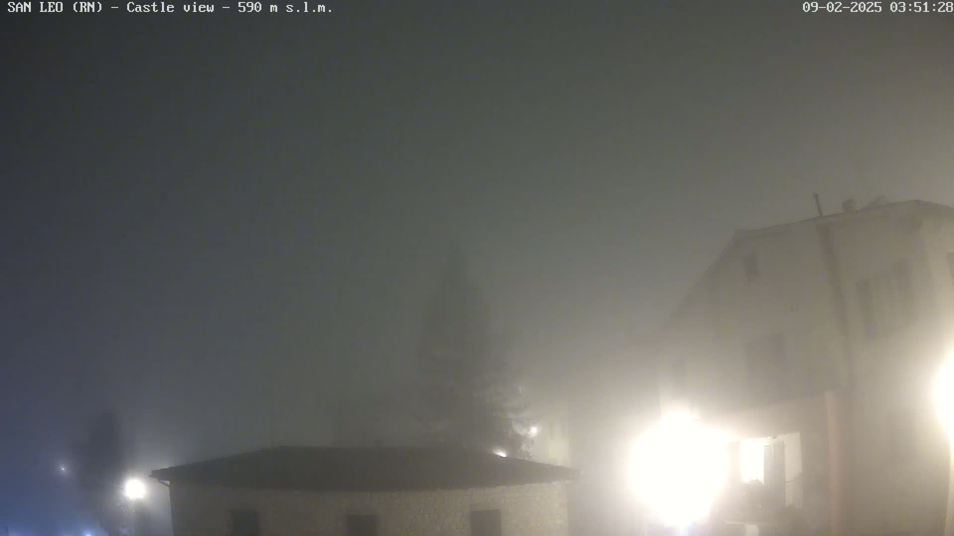 Two buildings are dimly lit by streetlights in thick fog at night.