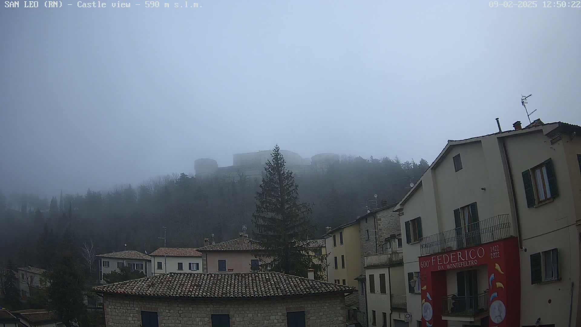 A foggy, overcast day reveals a hilltop castle partially obscured by mist, overlooking a village of stone buildings with terracotta roofs.