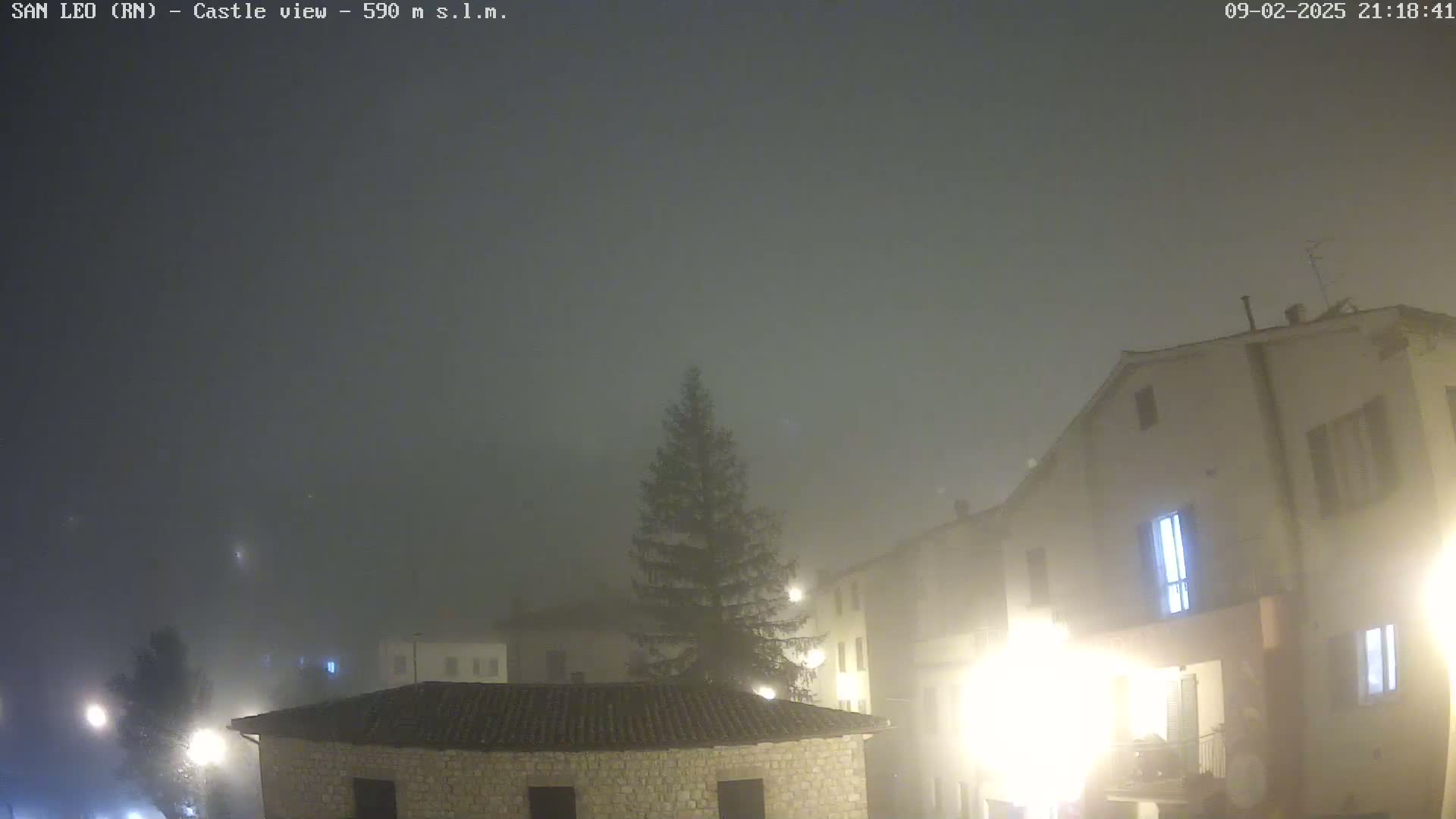 A foggy night scene shows a village square with stone buildings, a tall evergreen tree, and dimly lit streetlights.