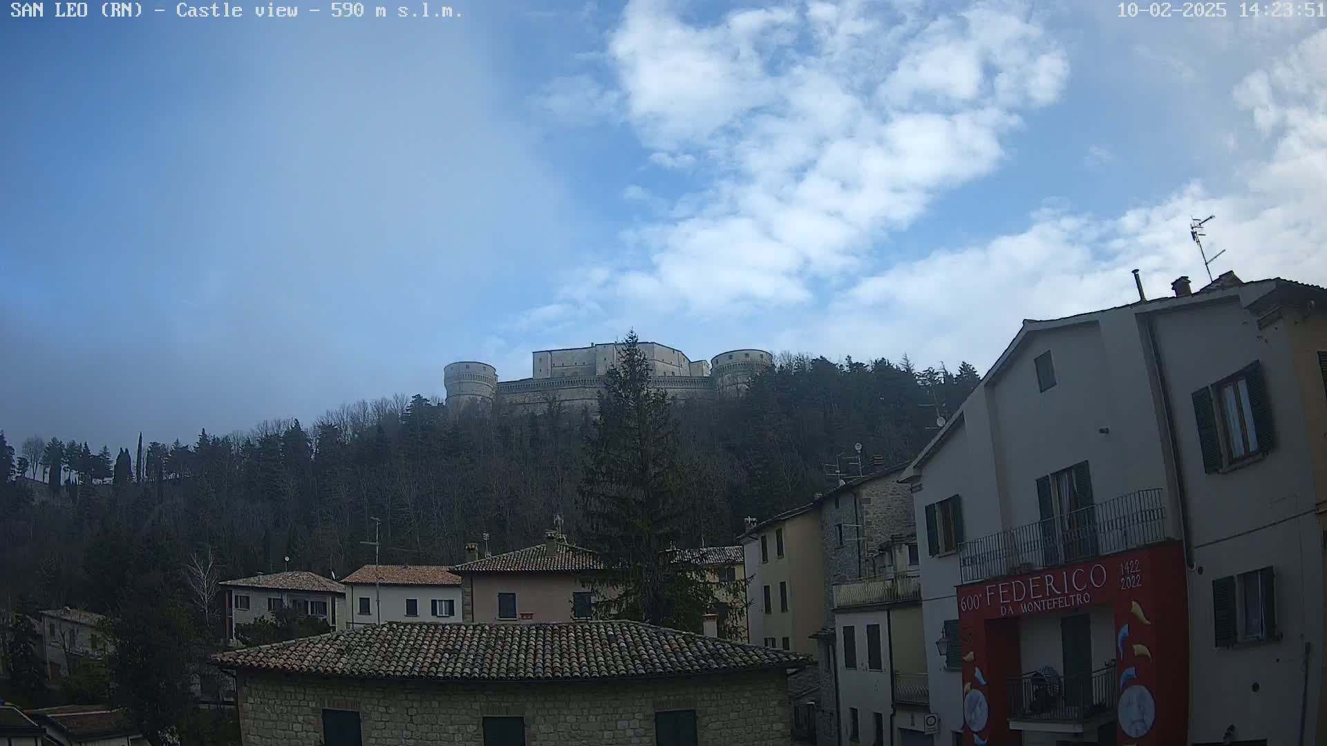 A large stone castle sits atop a hill covered in trees under a partly cloudy sky, with a village of stone buildings in the foreground.