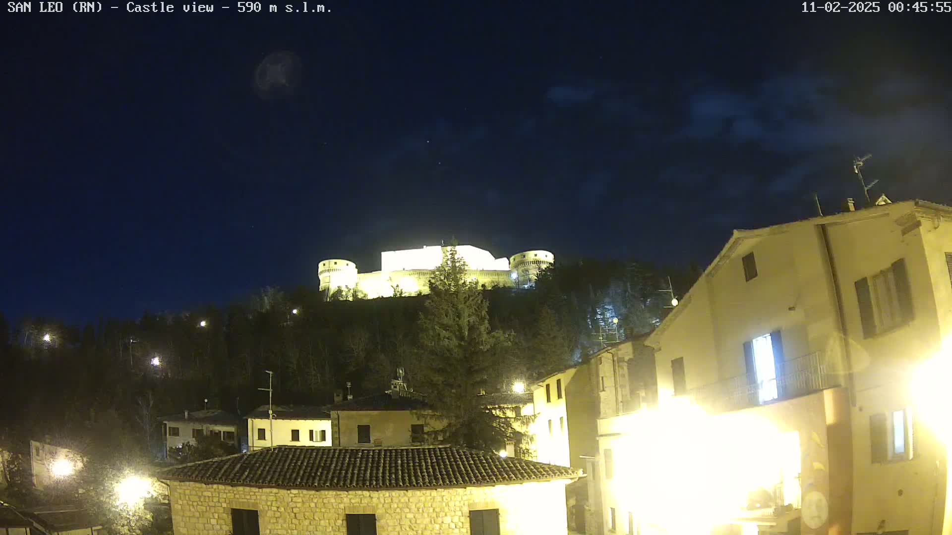 A nighttime view of a hilltop castle illuminated against a dark, slightly cloudy sky, overlooking a village with buildings lit by streetlights.
