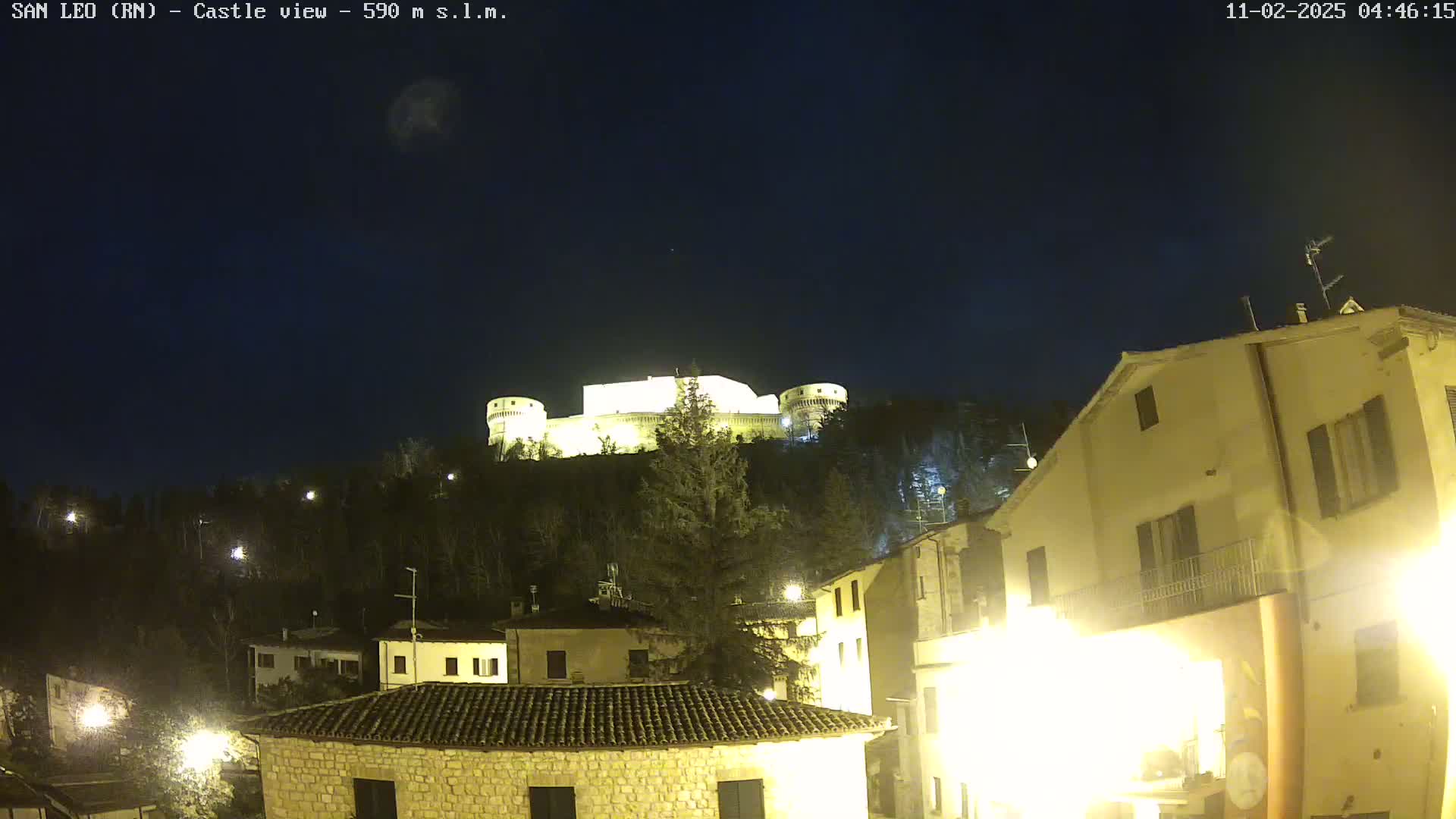 A nighttime view of a hilltop castle illuminated against a dark, clear sky, overlooking a village with buildings lit up below.