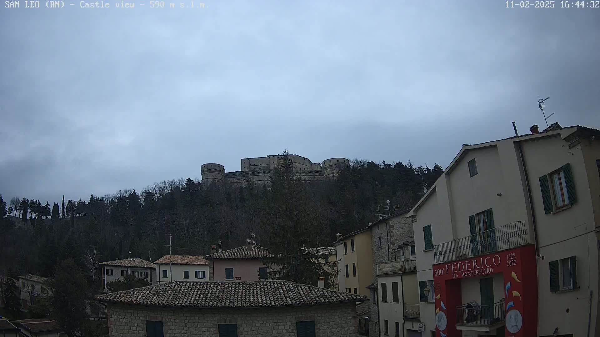 A stone castle sits atop a hill covered in leafless trees under a cloudy sky, overlooking a town of stone buildings with terracotta roofs.