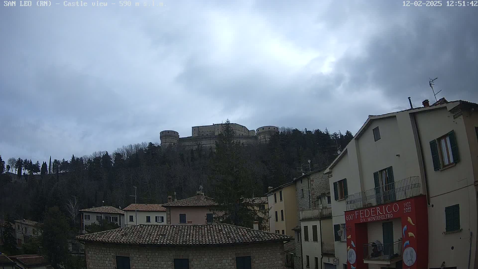 A stone castle sits atop a hill covered in leafless trees under an overcast sky, overlooking a village of stone buildings with terracotta roofs.