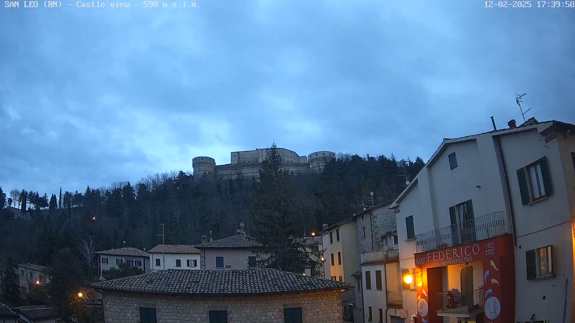 A stone castle sits atop a hill overlooking a town at twilight under a cloudy sky.