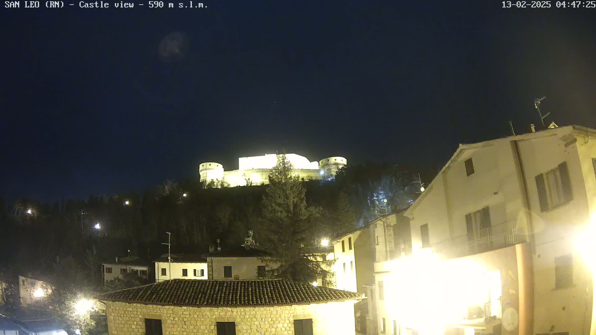 A brightly lit castle sits atop a hill overlooking a town at night under a clear, dark sky.