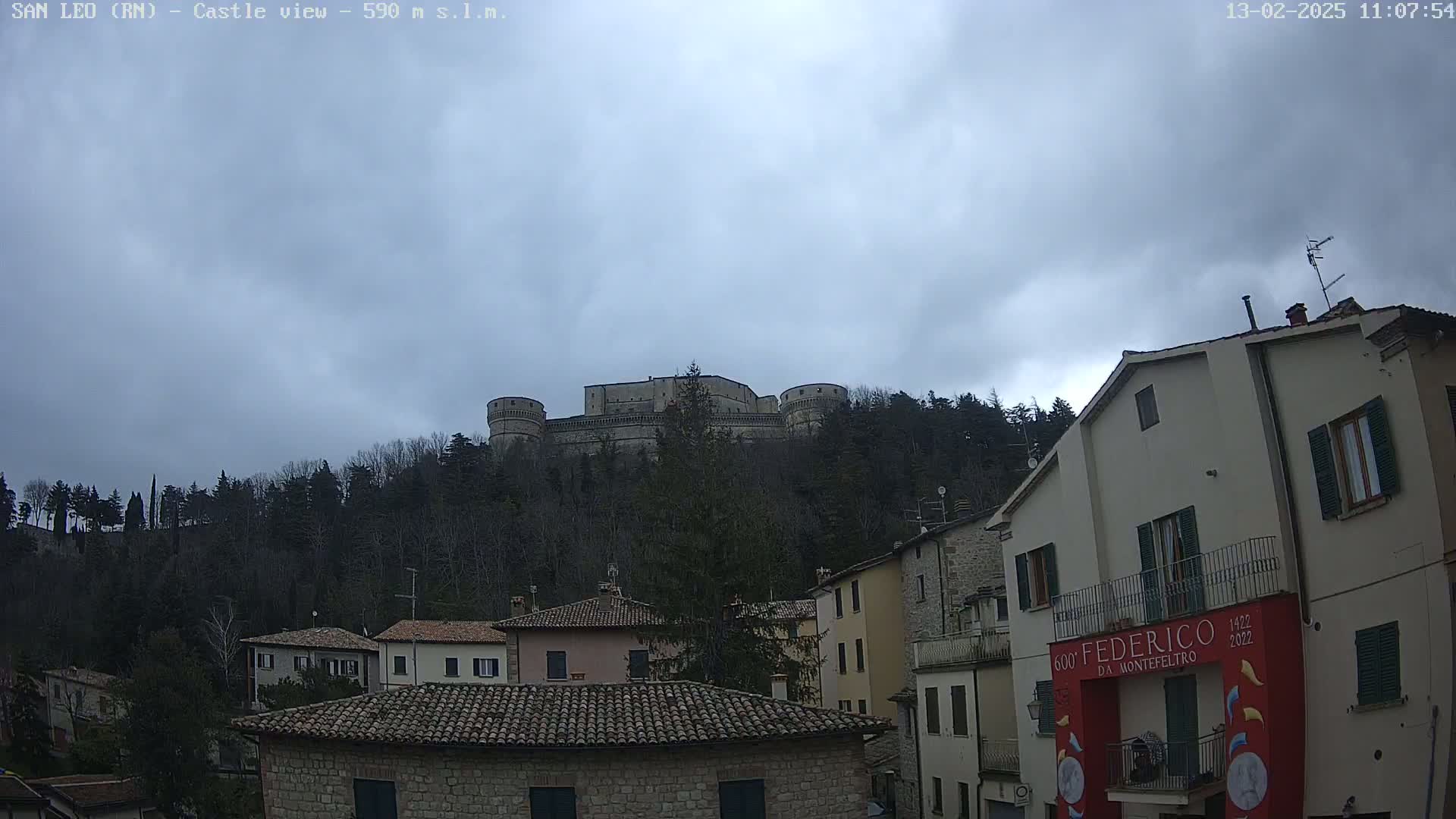 A stone castle sits atop a hill overlooking a town of low, stone buildings under a cloudy sky.