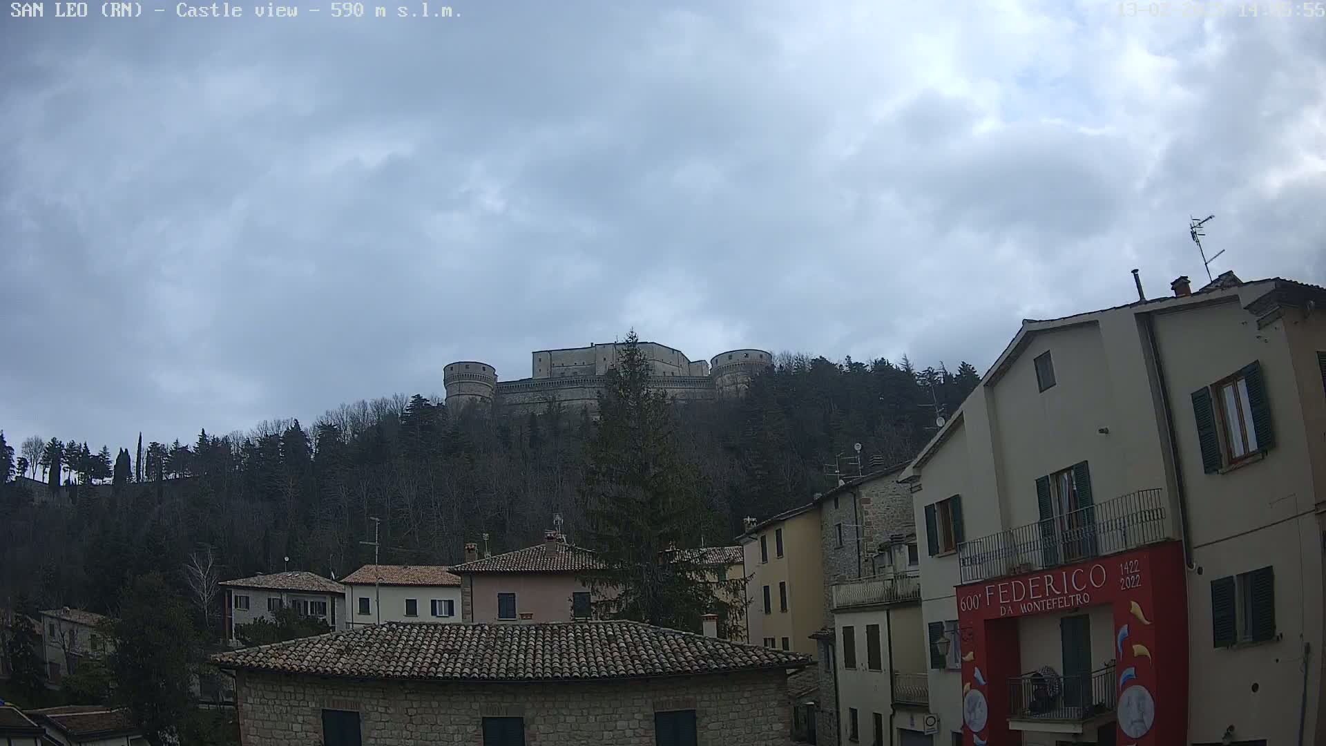 A large stone castle sits atop a hill covered in leafless trees under a cloudy sky, overlooking a town of low stone buildings.