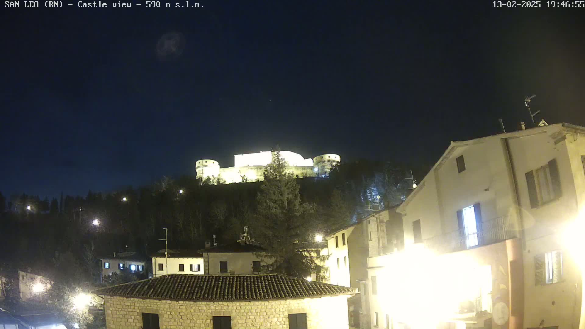 A brightly lit castle sits atop a hill at night, overlooking a town with stone buildings under a clear, dark sky.