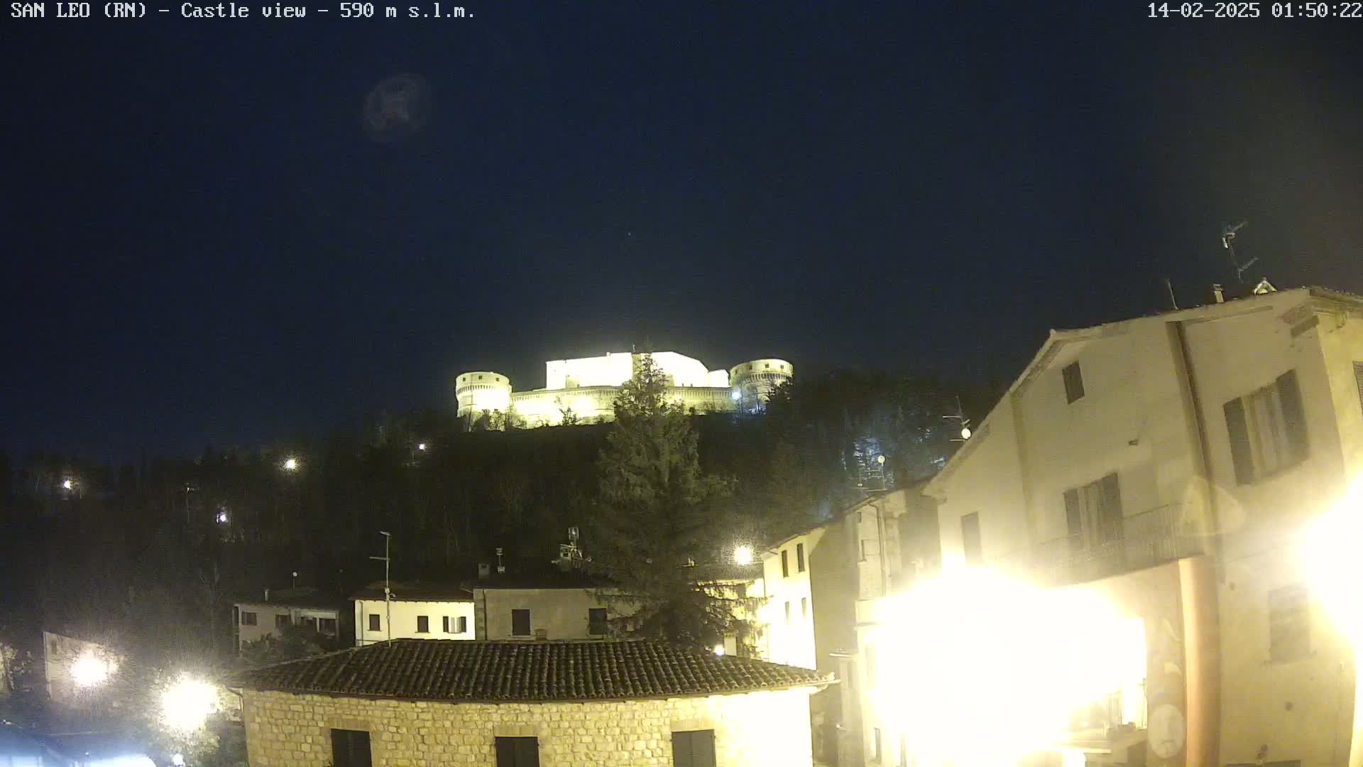 A brightly lit castle sits atop a hill at night, overlooking a town with illuminated buildings.