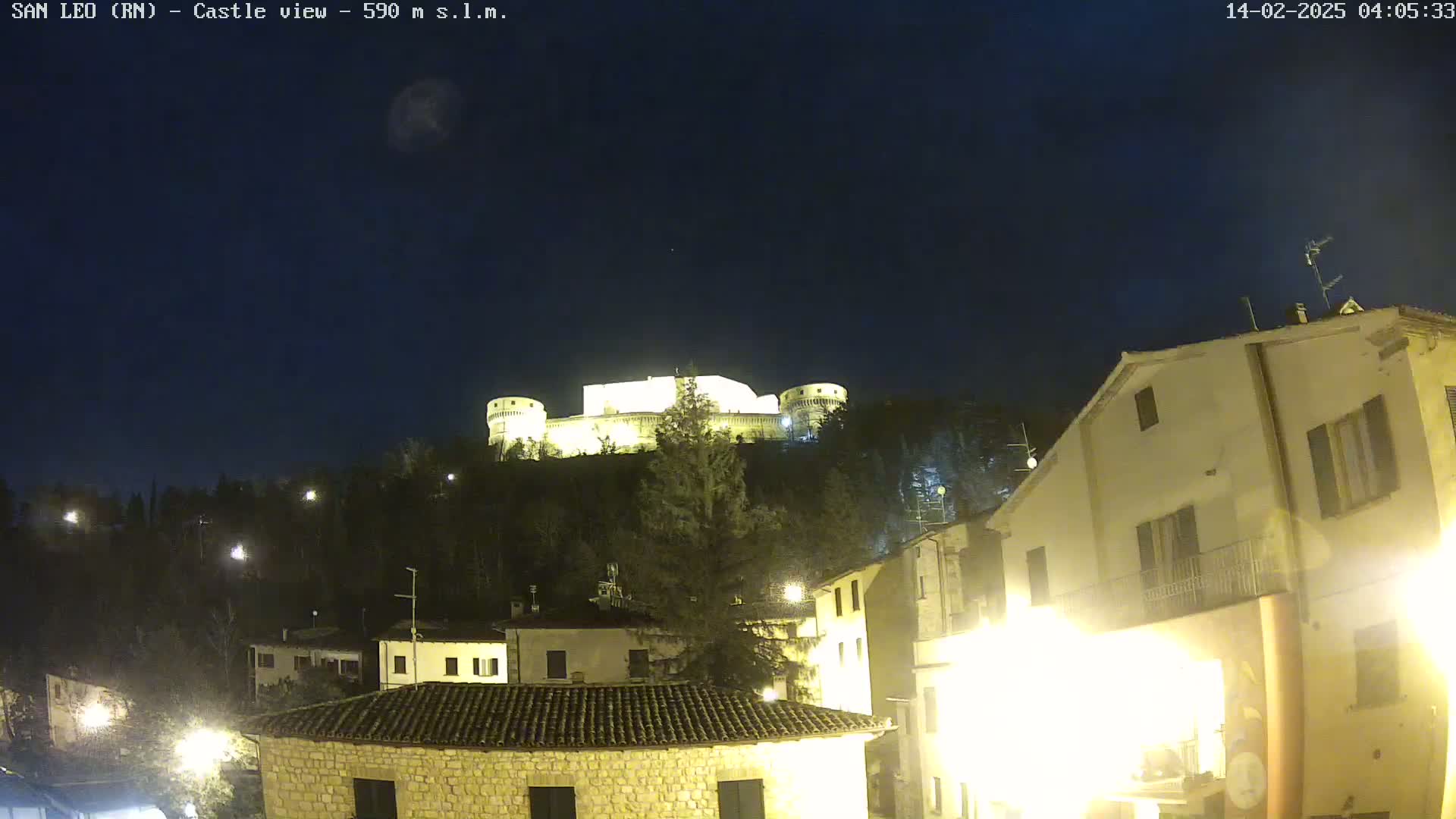 A nighttime view of a hilltop castle illuminated against a clear, dark sky, with a town's buildings and streetlights visible in the foreground.