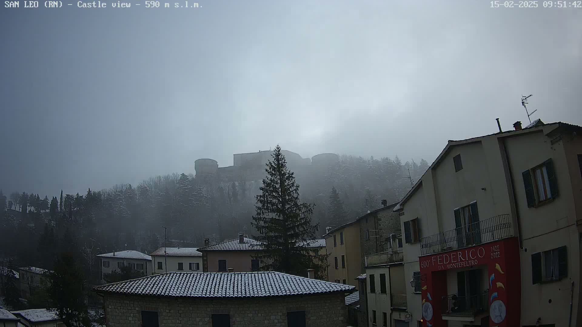 A snow-dusted castle sits atop a foggy hill overlooking a village of snow-covered rooftops.