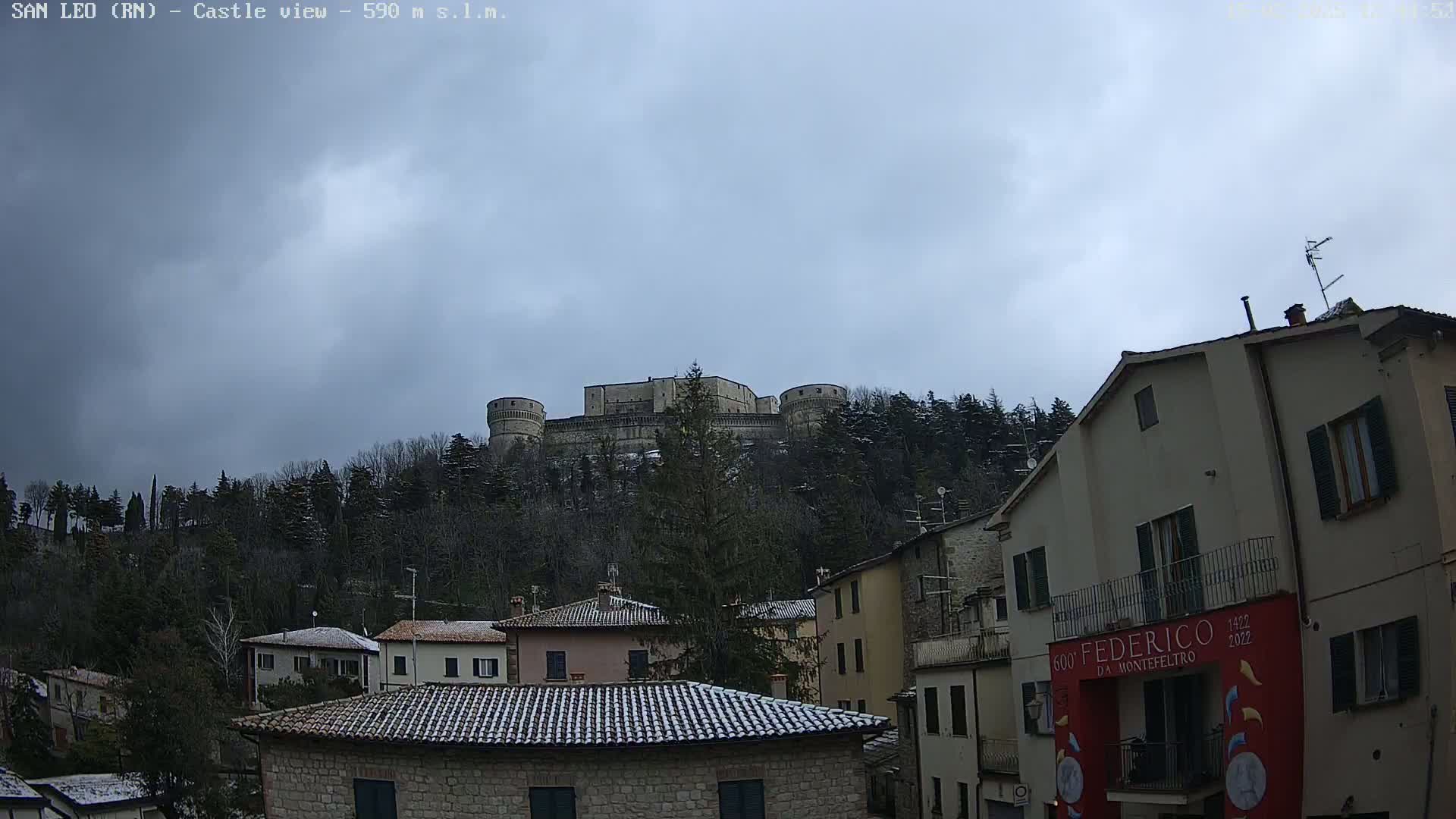 A large stone castle sits atop a snow-dusted hill behind a town of buildings with gray roofs, under a cloudy sky.