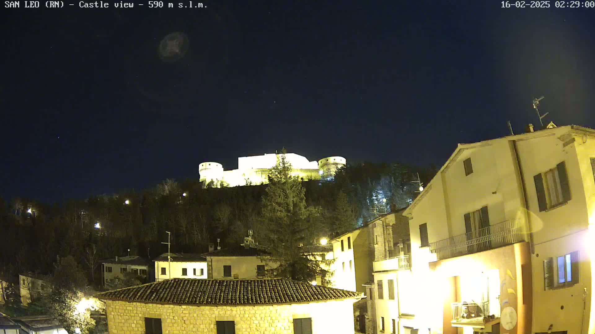 A nighttime view of a hilltop castle illuminated against a dark, clear sky overlooking a village with buildings lit up below.