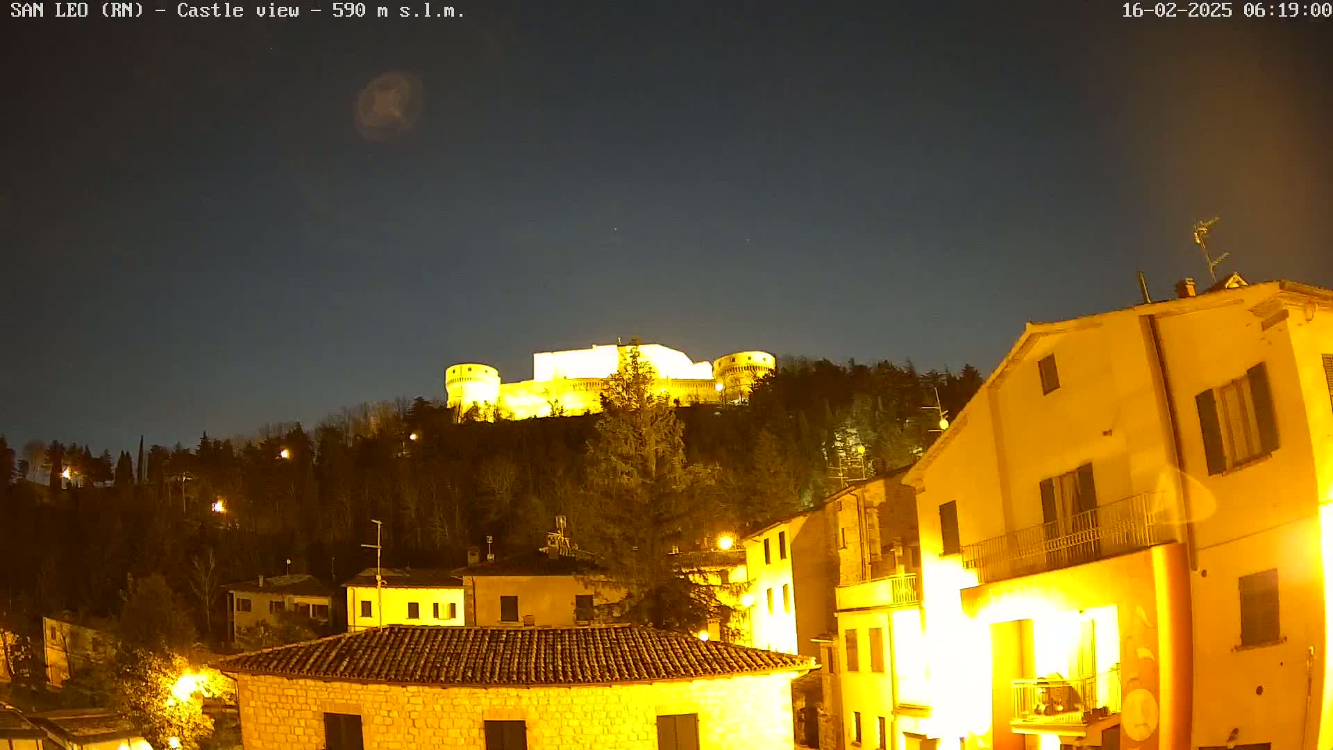 A brightly lit castle sits atop a hill at night, overlooking a town of illuminated buildings under a dark, clear sky.