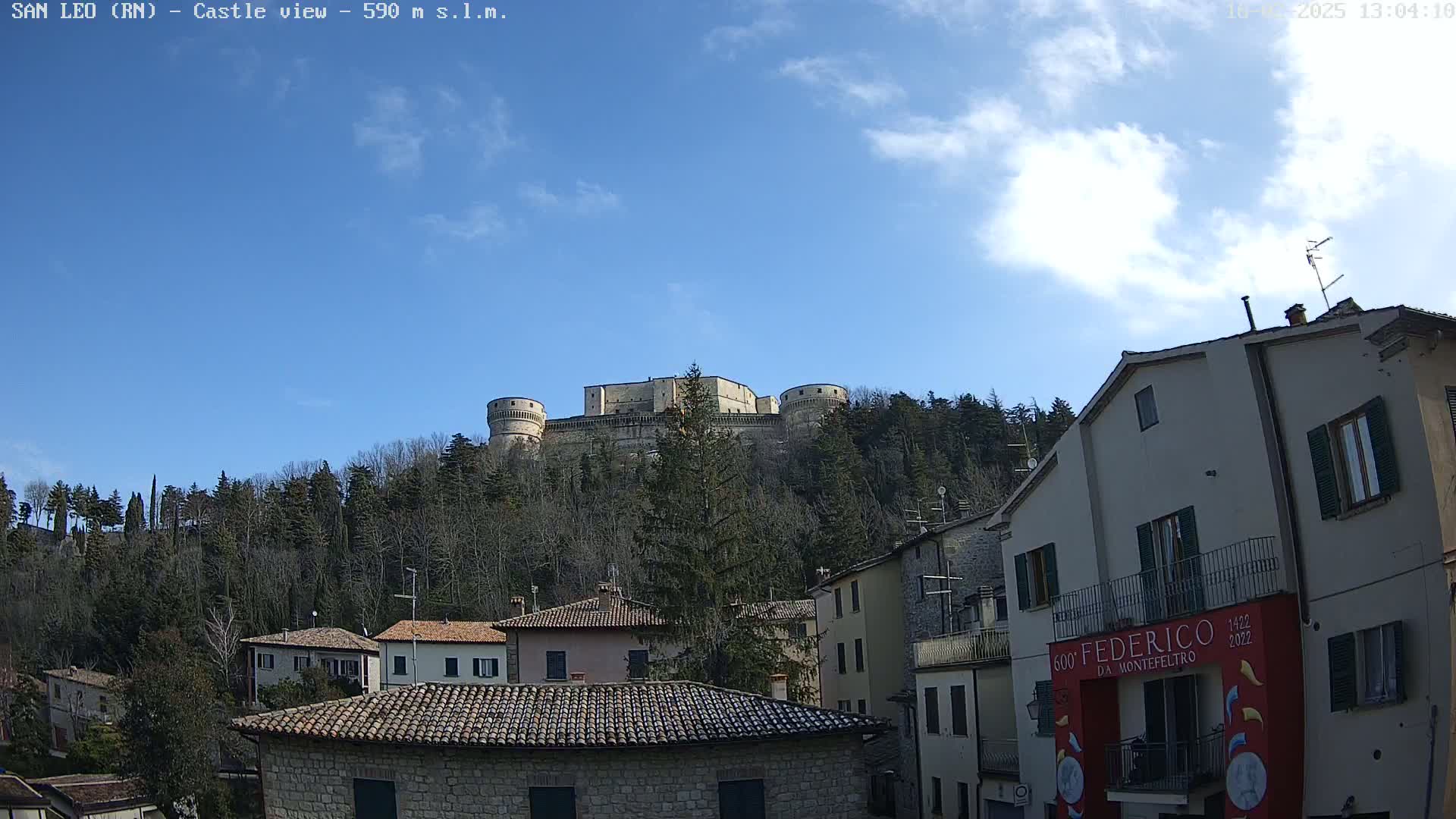 A stone castle sits atop a hill covered in leafless trees under a mostly clear blue sky, overlooking a village of stone buildings.