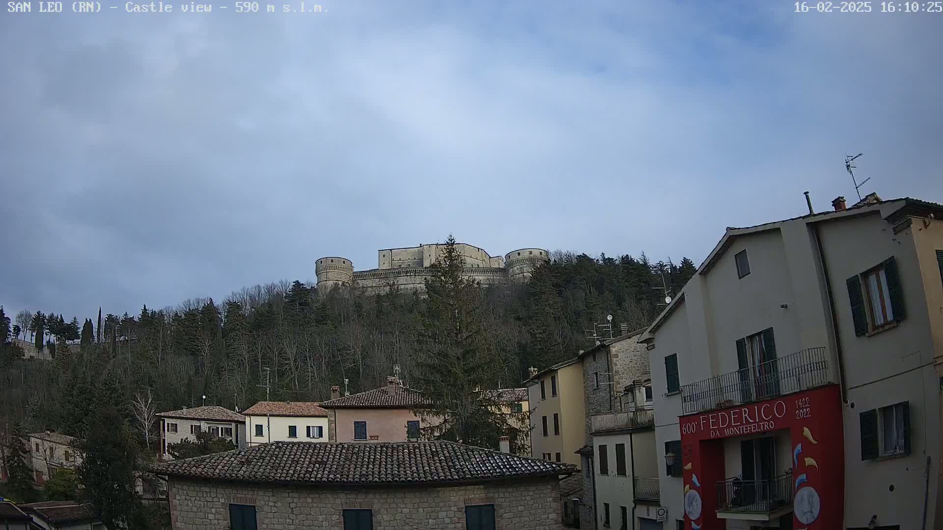 A large stone castle sits atop a hill overlooking a town of low stone buildings under a mostly cloudy sky.