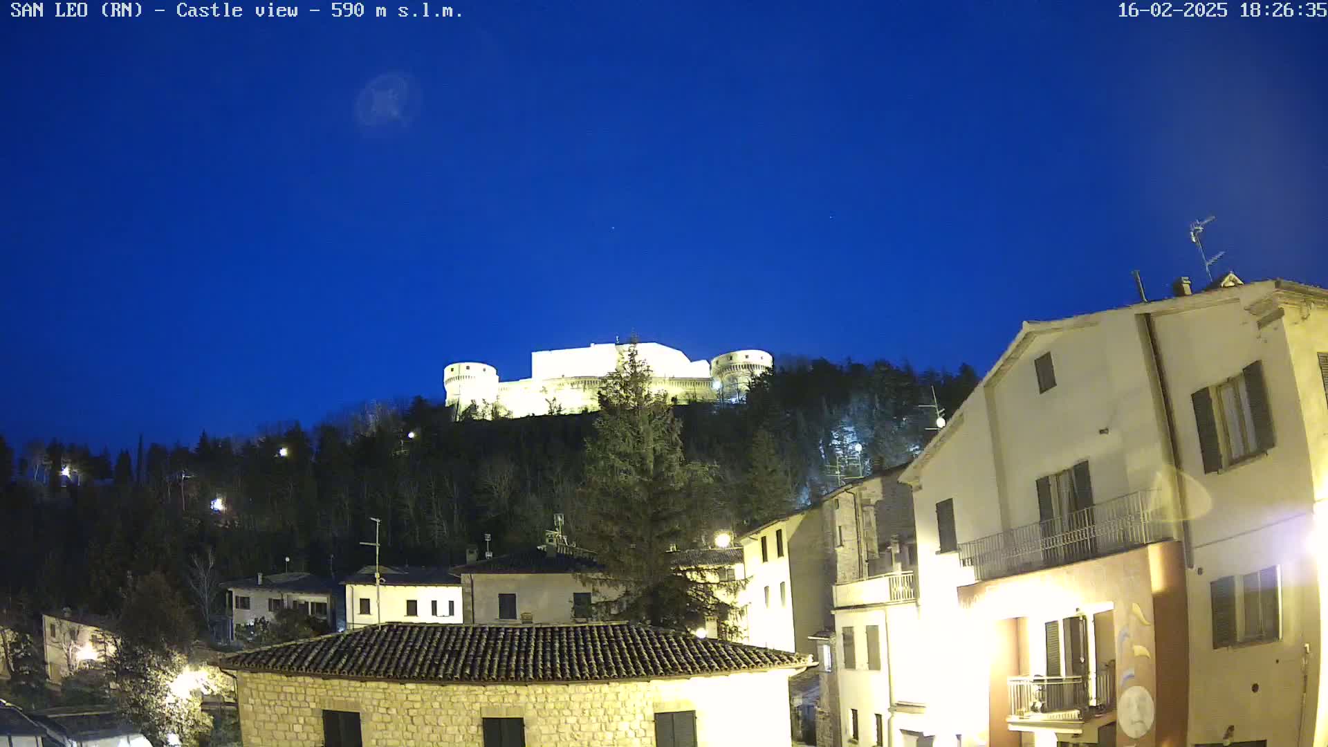 A brightly lit castle sits atop a hill overlooking a town at night under a clear, dark blue sky.