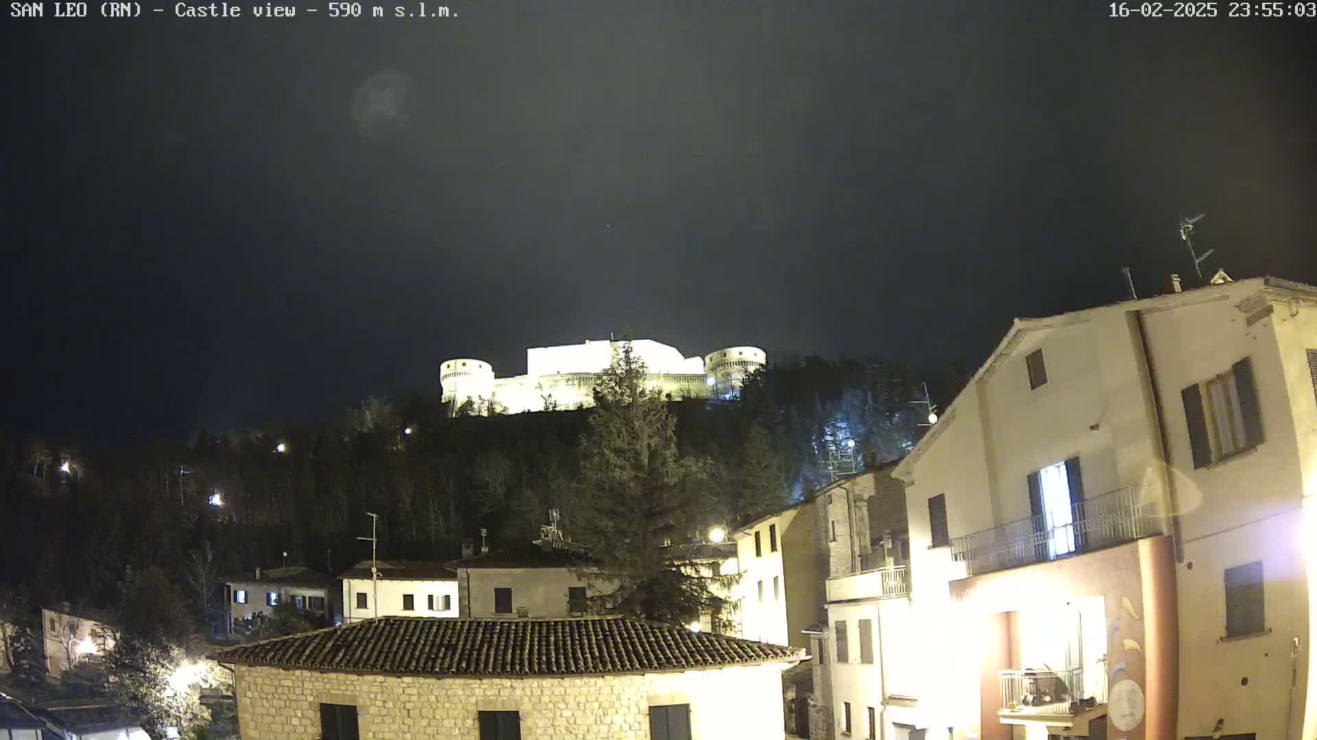 A nighttime view of a town with a brightly lit castle perched atop a hill in the background under a dark, cloudy sky.