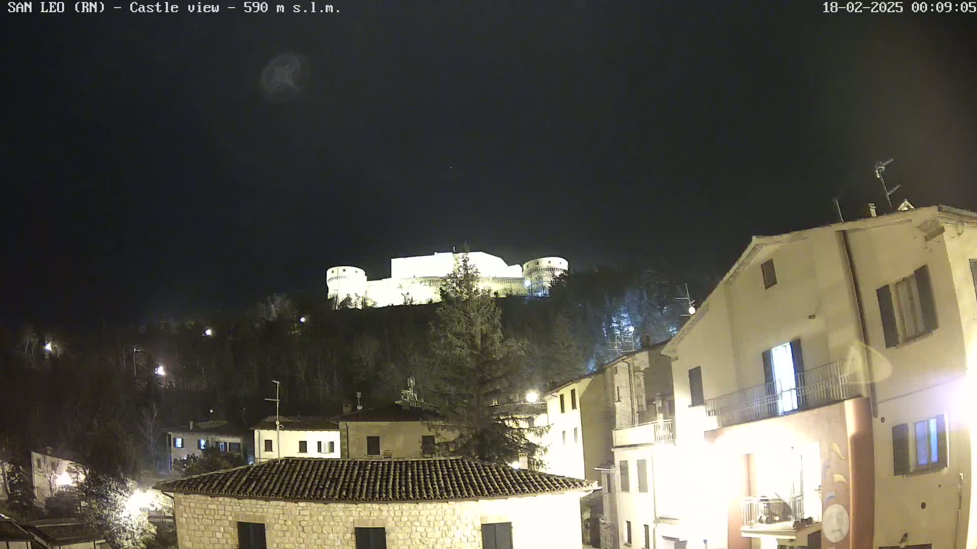 A nighttime view of a hilltop castle illuminated against a dark sky, overlooking a village with buildings lit up.