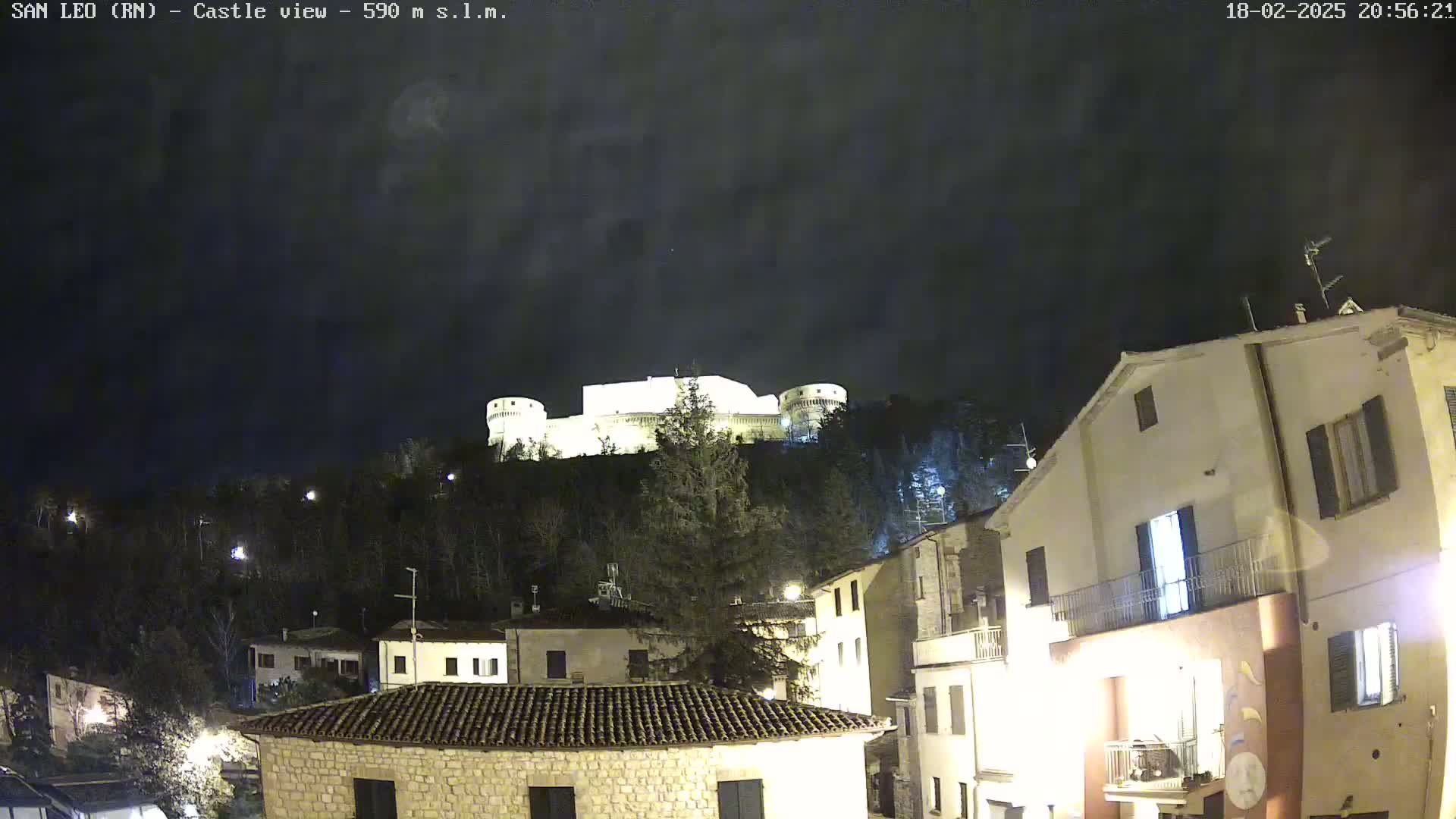 A nighttime view of a hilltop castle overlooking a town with buildings illuminated under a dark, cloudy sky.
