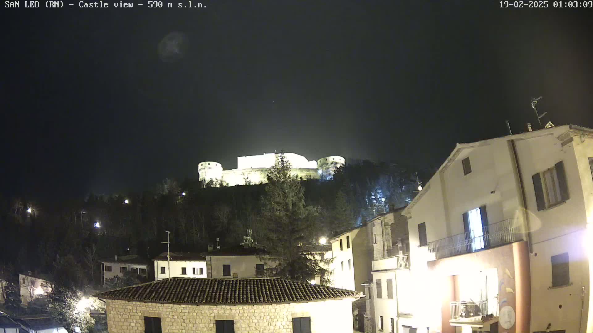 A nighttime view of a hilltop castle illuminated against a dark, clear sky, overlooking a village with several buildings lit up.