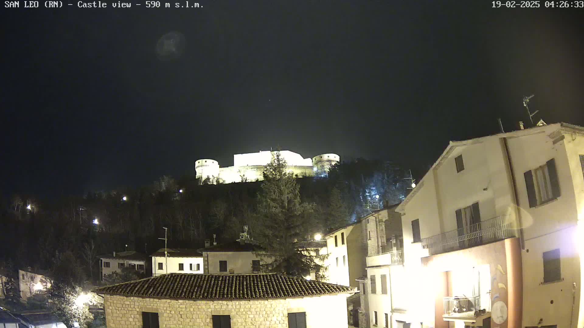 A nighttime view of a hilltop castle illuminated against a dark, clear sky, overlooking a village with buildings lit up below.