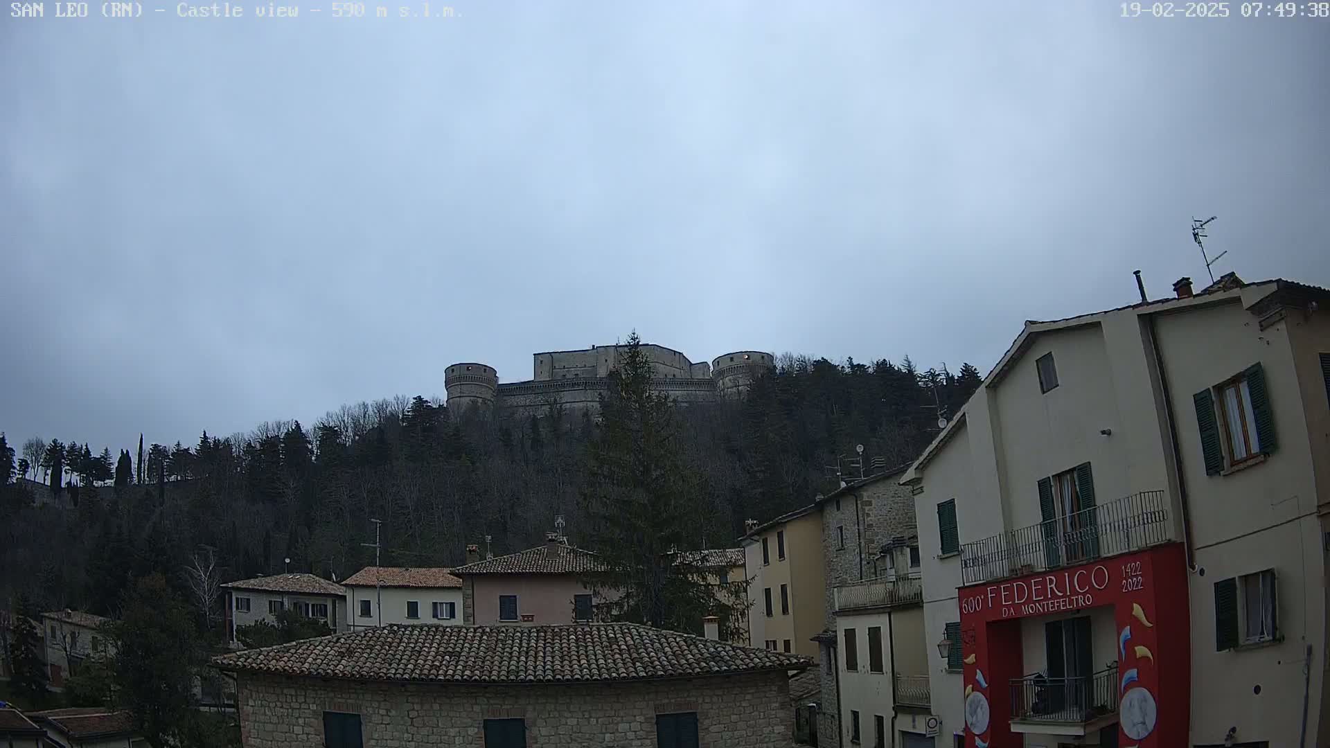A large stone castle sits atop a hill overlooking a town of low buildings under a cloudy sky.