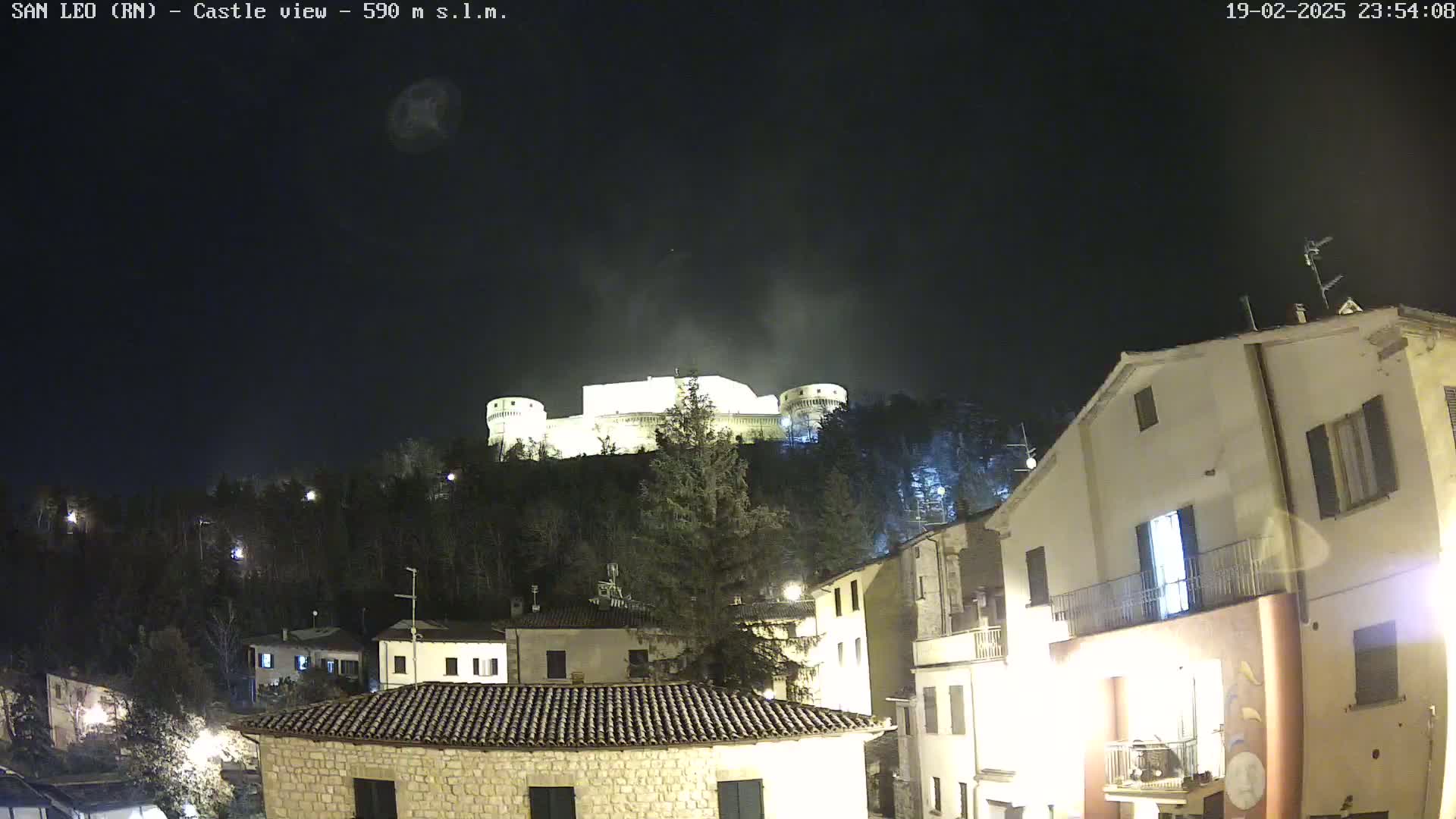 A nighttime view of a hilltop castle illuminated against a dark, clear sky, overlooking a village with buildings lit up below.