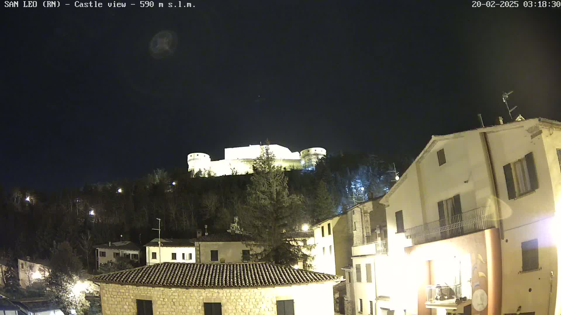 A nighttime view of a hilltop castle illuminated against a dark, clear sky, overlooking a town with buildings lit up below.