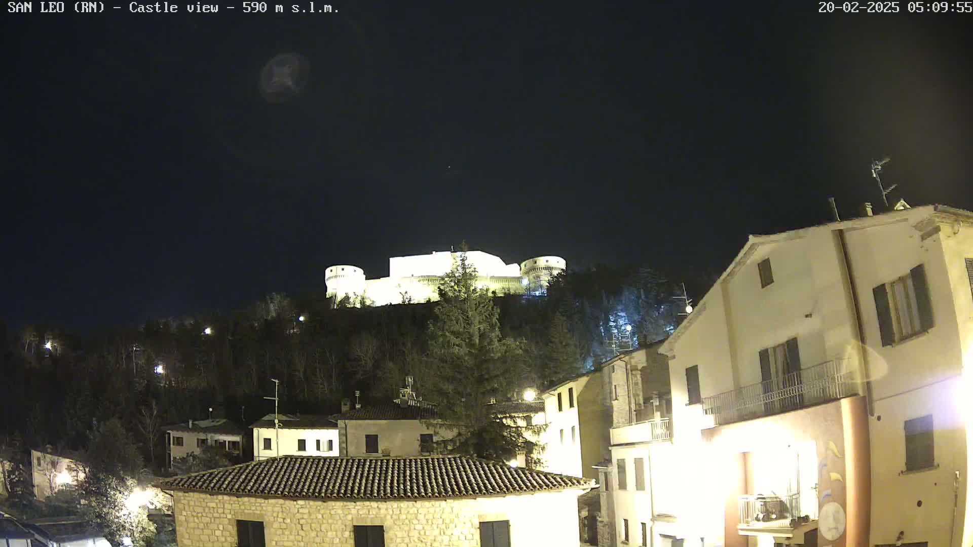 A nighttime view of a hilltop castle illuminated against a dark sky, overlooking a town with buildings lit up below.