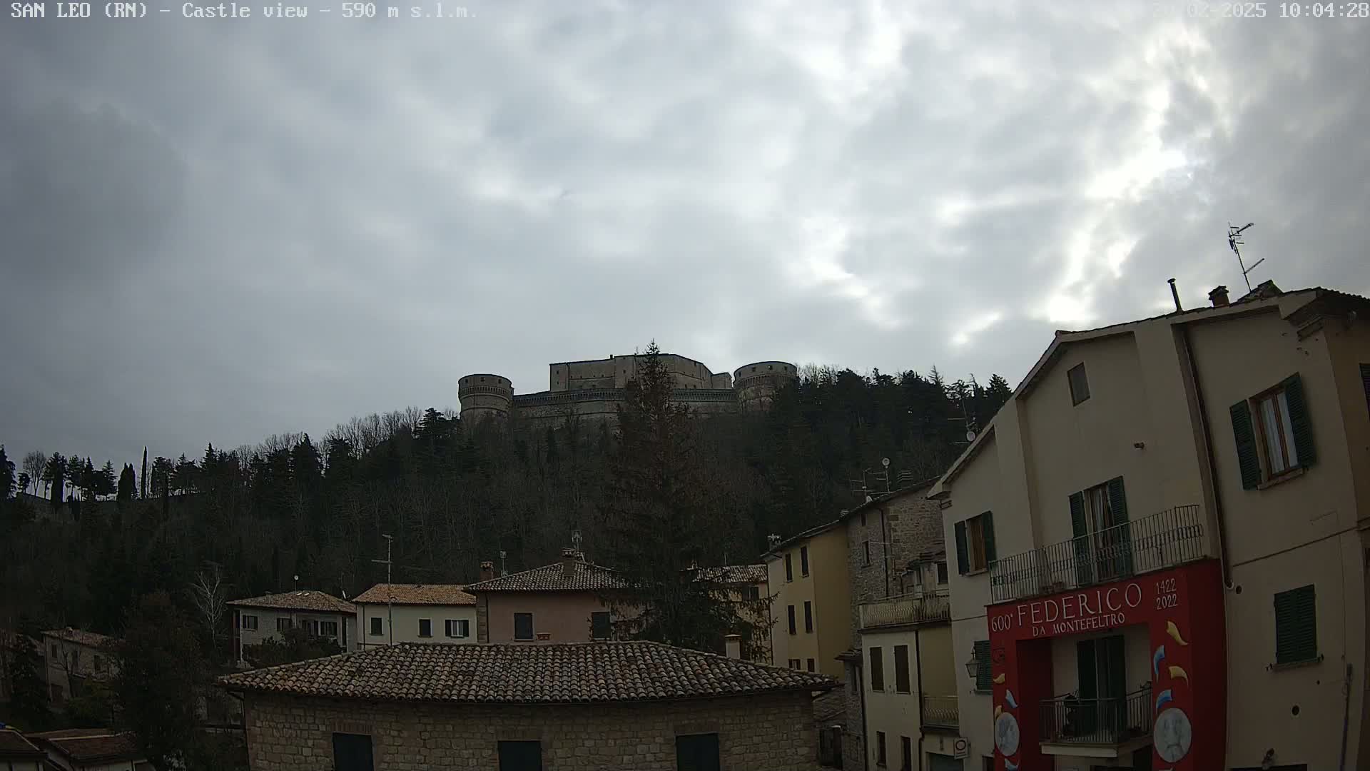 A stone castle sits atop a hill covered in dark trees under a cloudy sky, overlooking a town with low buildings and terracotta roofs.