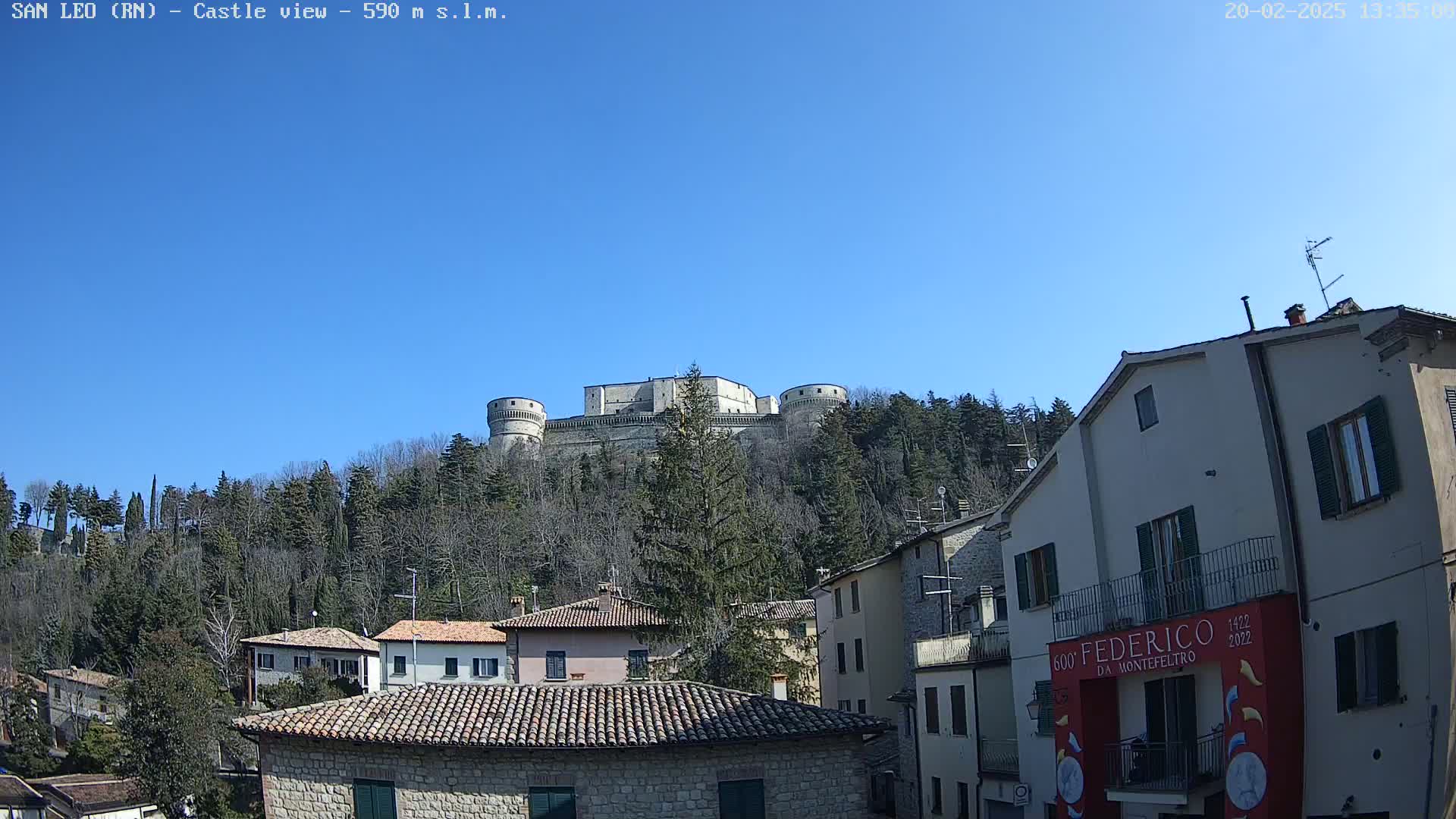 A large stone castle sits atop a hill covered in leafless trees under a clear blue sky, overlooking a town of stone buildings with terracotta roofs.
