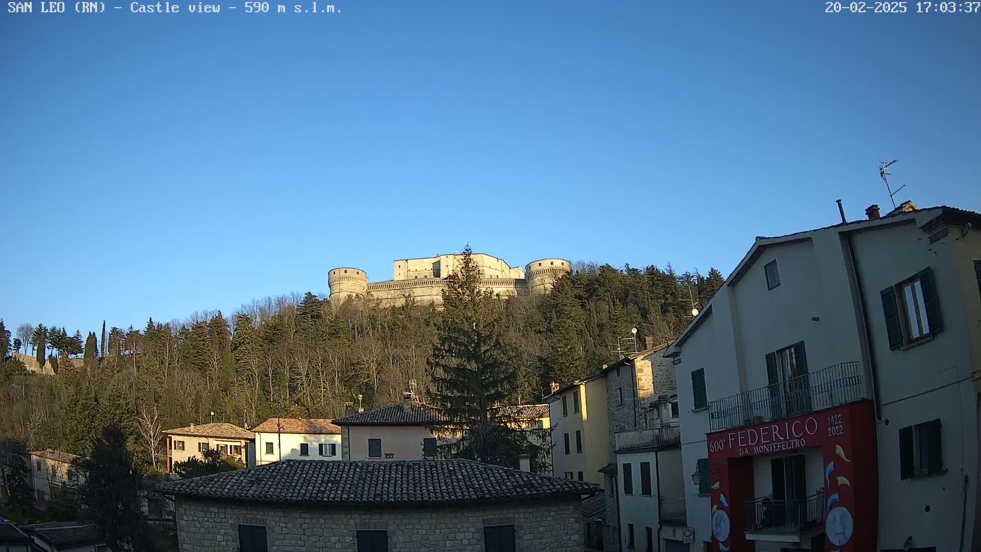 A large stone castle sits atop a tree-covered hill under a clear blue sky, overlooking a village of stone buildings.
