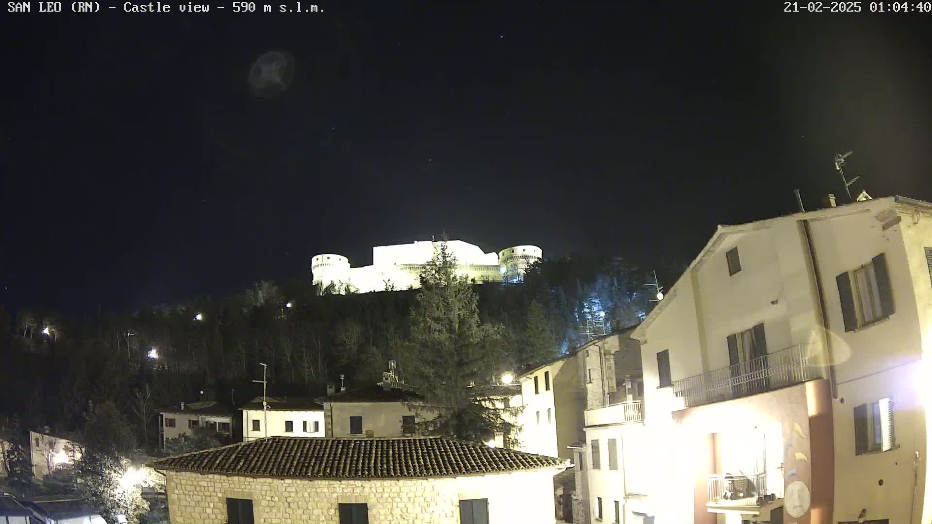 A nighttime view of a hilltop castle illuminated against a dark, clear sky, overlooking a village with buildings lit by streetlights.