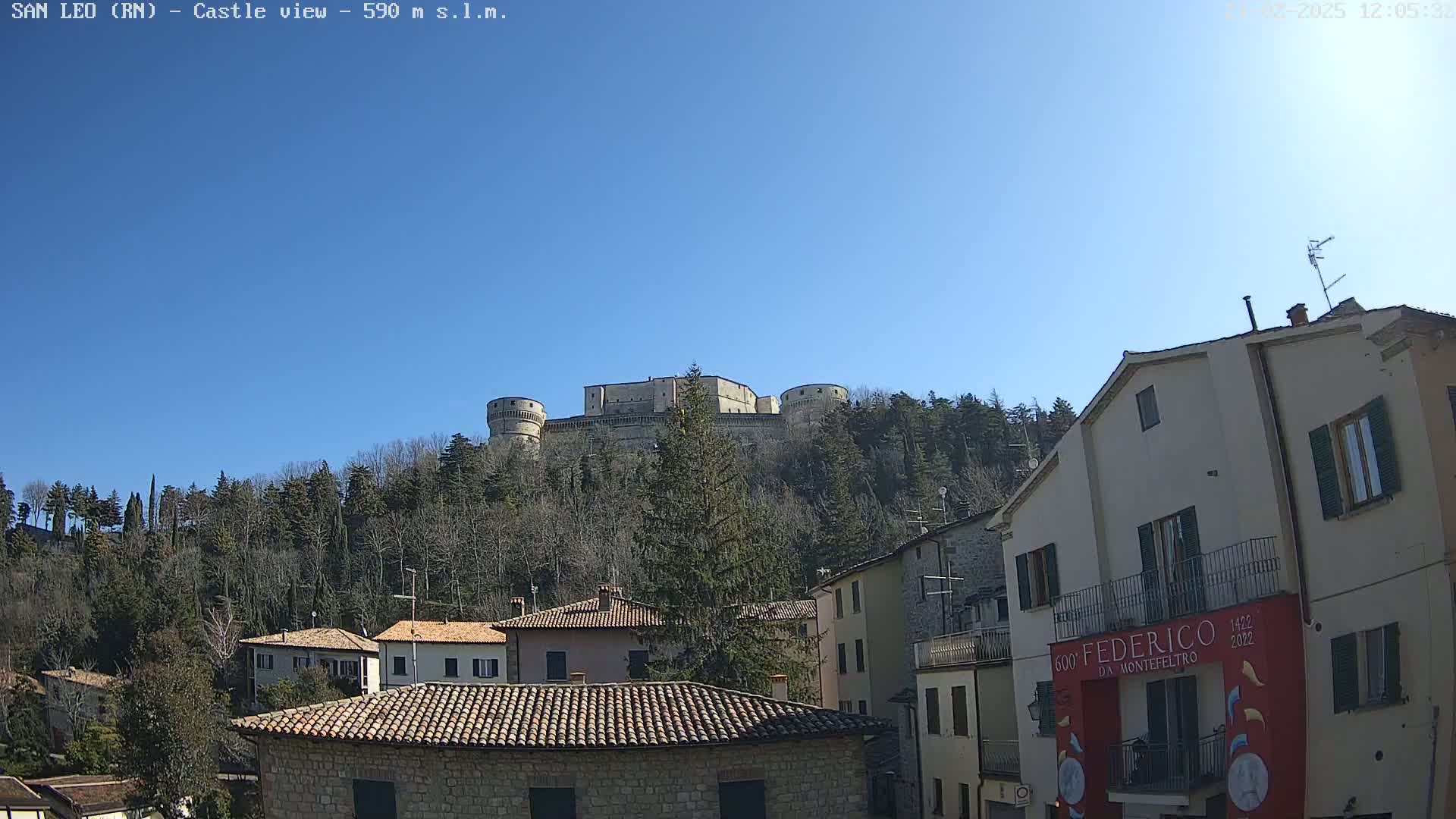 A large stone castle sits atop a hill overlooking a town of low stone buildings under a clear blue sky.