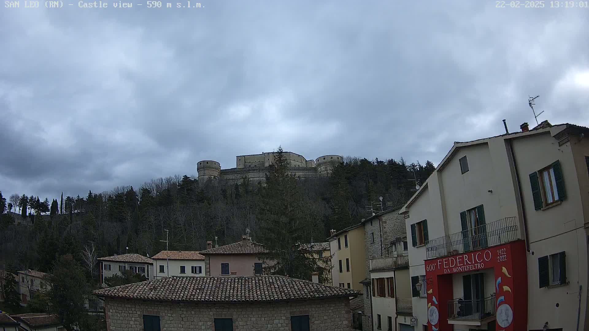A large stone castle sits atop a hill covered in leafless trees under a cloudy sky, overlooking a town of closely spaced buildings with terracotta roofs.