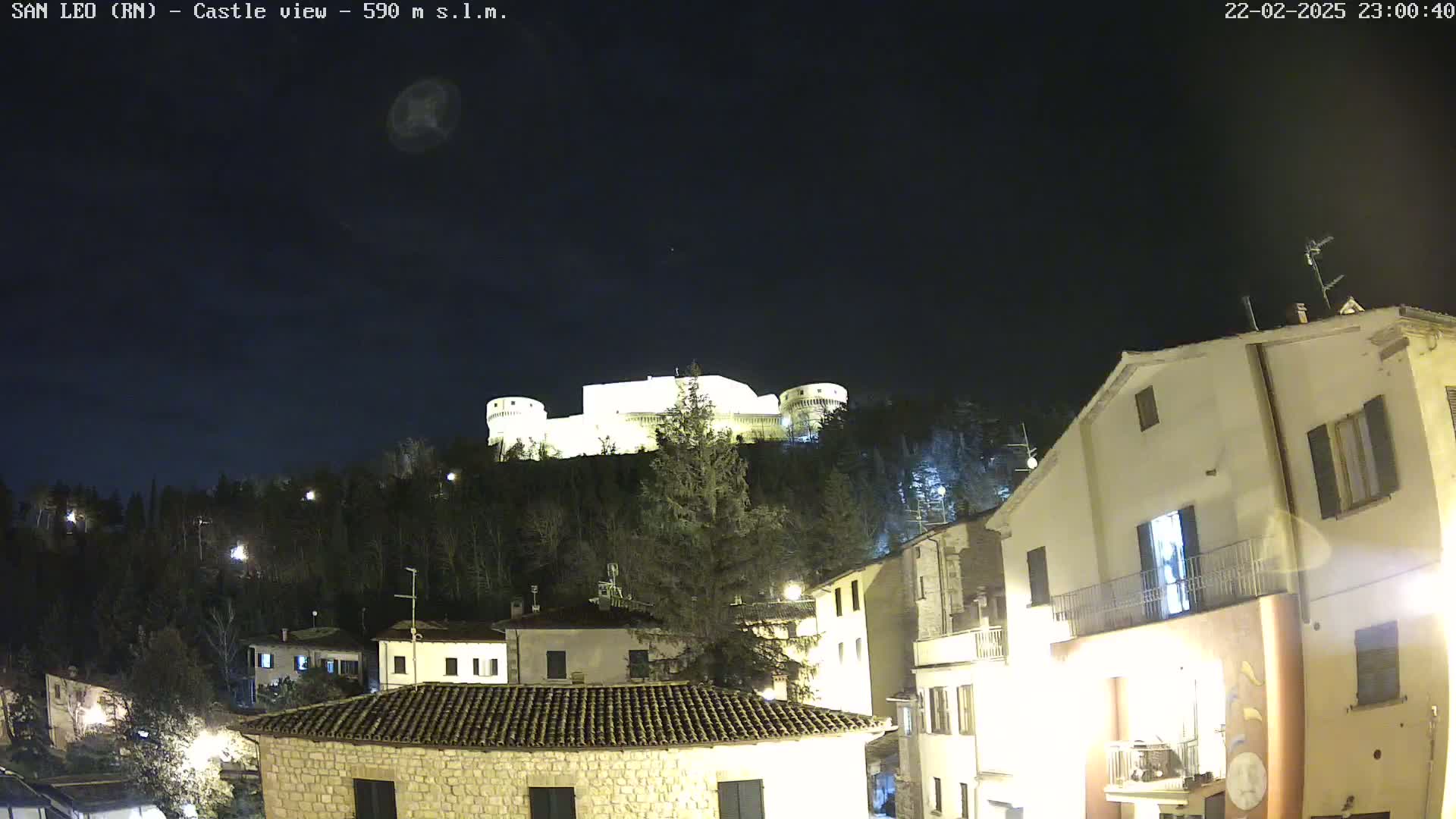 A nighttime view of a hilltop castle illuminated against a dark sky, overlooking a small town with buildings lit up.