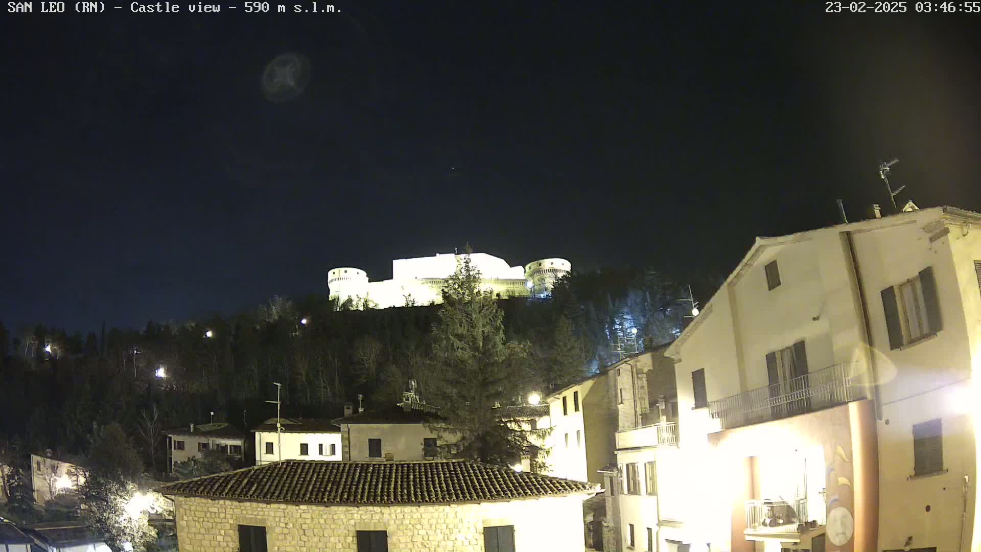 A brightly lit castle sits atop a hill at night, overlooking a town with several buildings illuminated under a clear, dark sky.