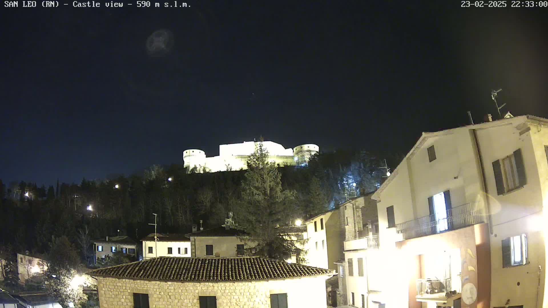 A nighttime view of a hilltop castle illuminated against a dark, clear sky, overlooking a village with several buildings.