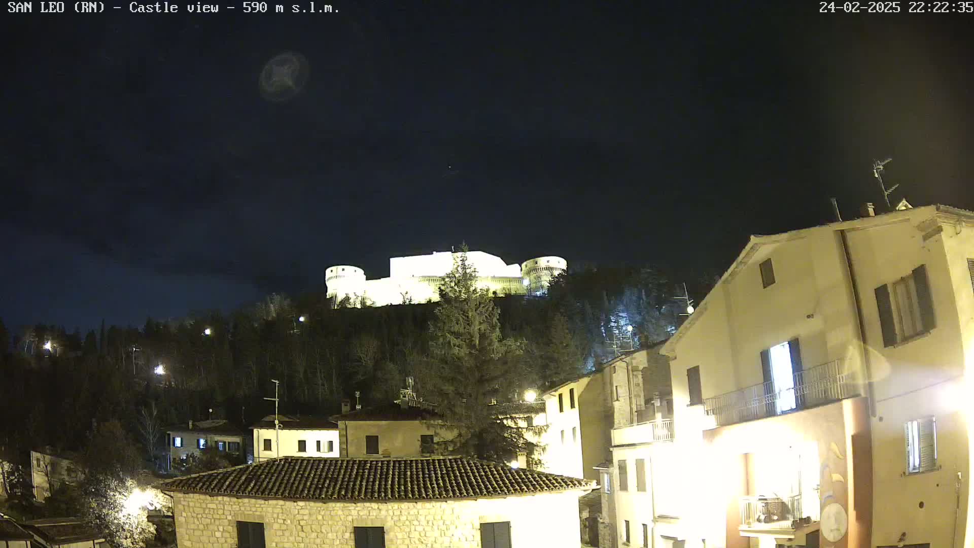 A nighttime view of a hilltop castle illuminated against a dark, clear sky, overlooking a village with lit buildings.