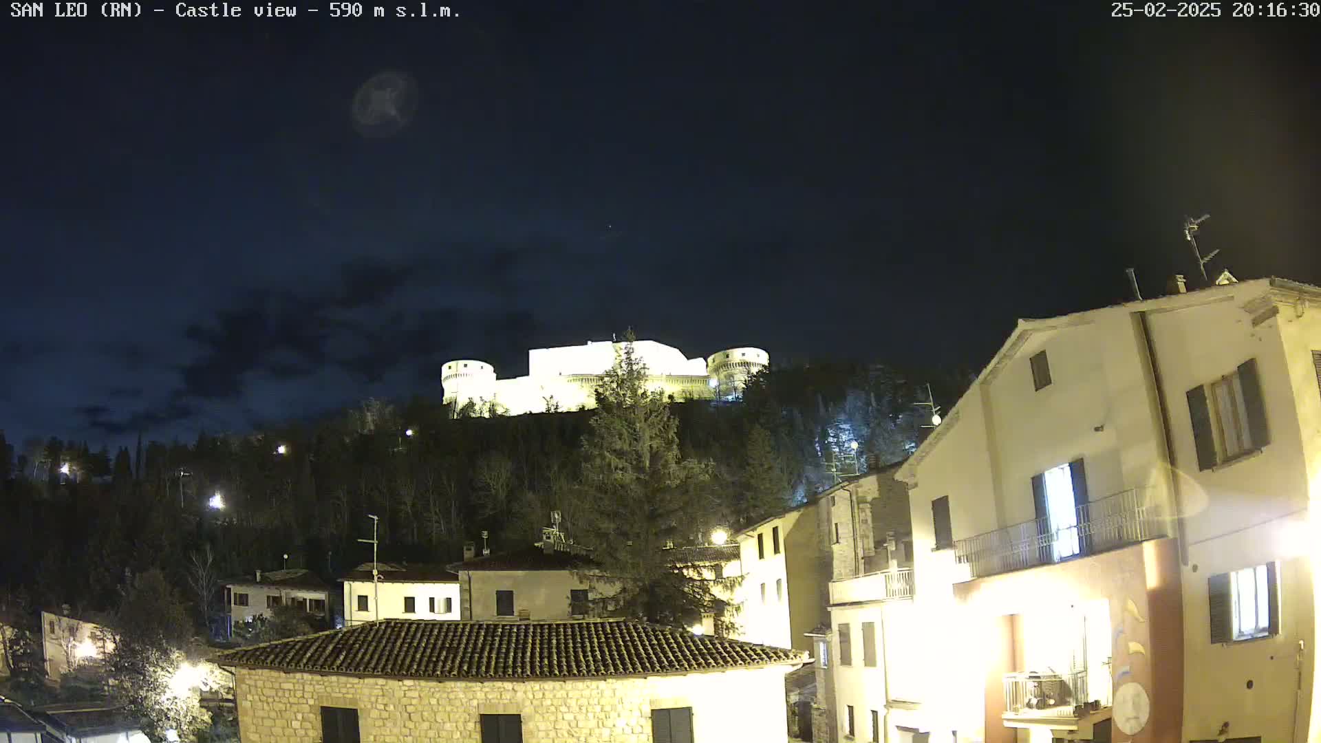 A nighttime view of a hilltop castle illuminated against a dark, cloudy sky, with a village of buildings nestled below.