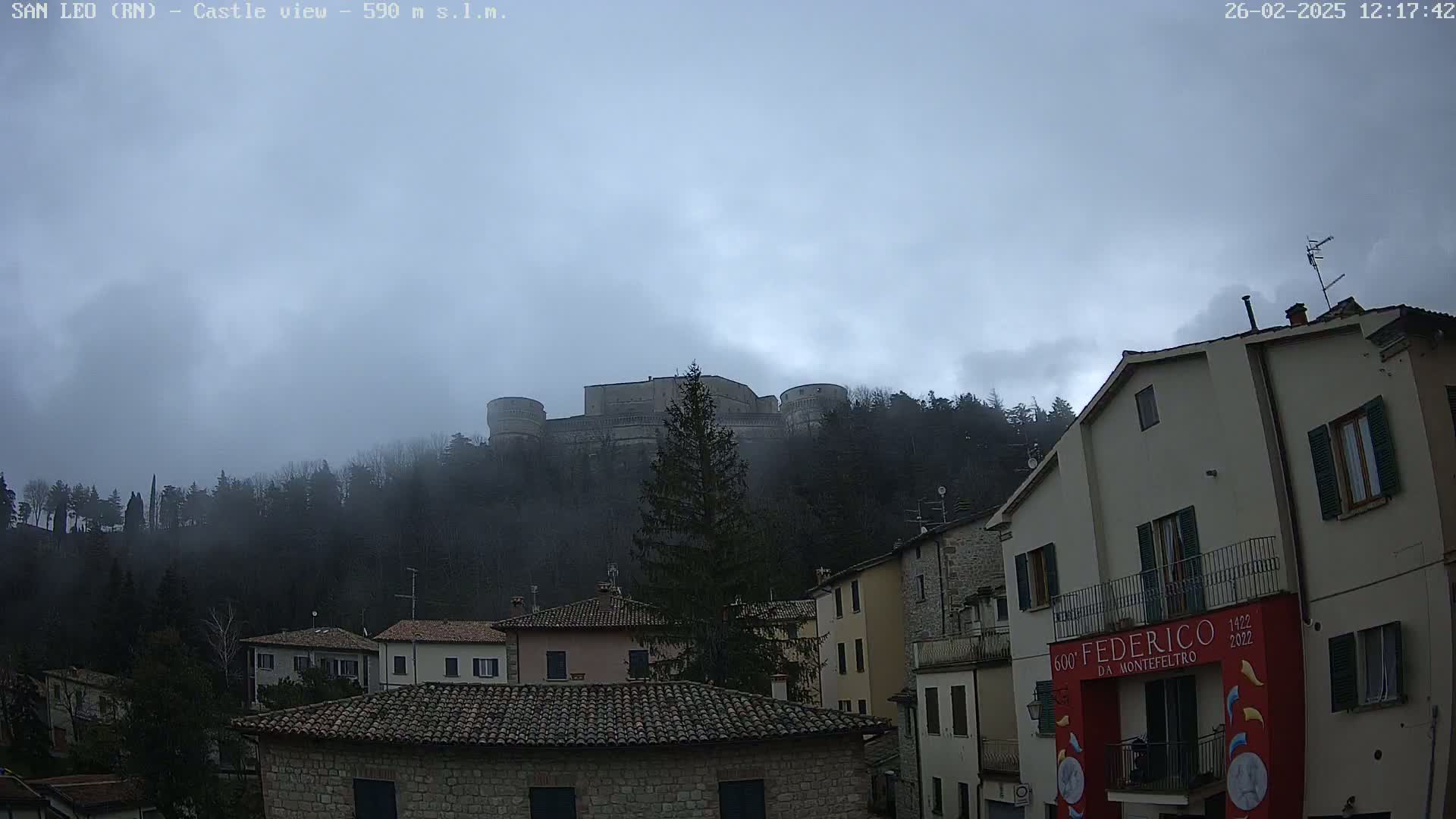 A stone castle sits atop a fog-covered hill behind a row of buildings in a town under an overcast sky.